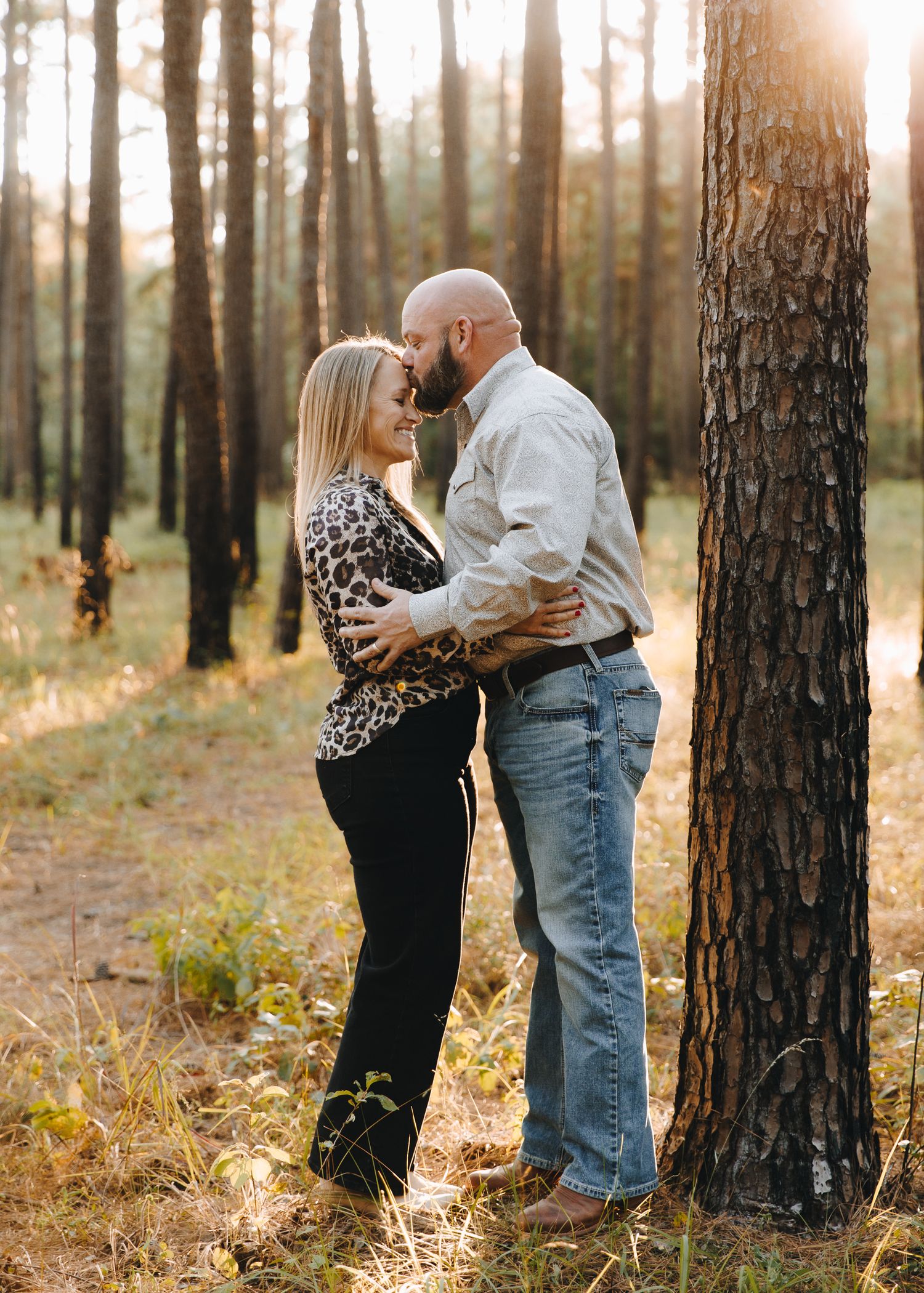 A couple shares a romantic moment leaning close together beside a tall pine tree at golden hour.