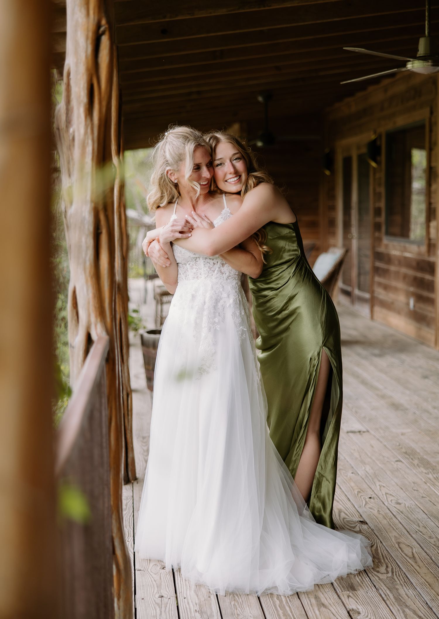 Bride in white lace gown hugging bridesmaid in olive green satin dress on wooden porch.