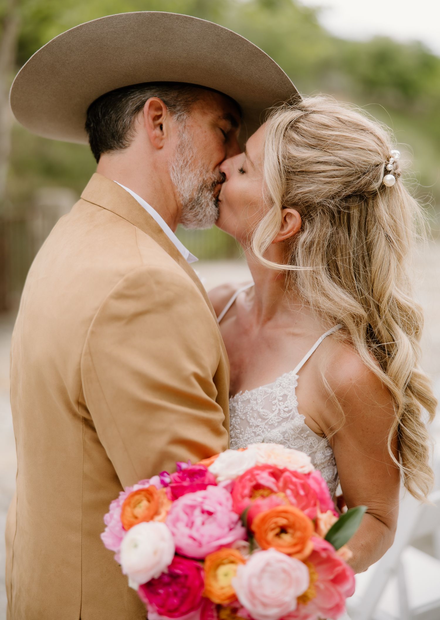 Bride and groom sharing a kiss, groom wearing a cowboy hat, bride holding a vibrant pink and orange bouquet.