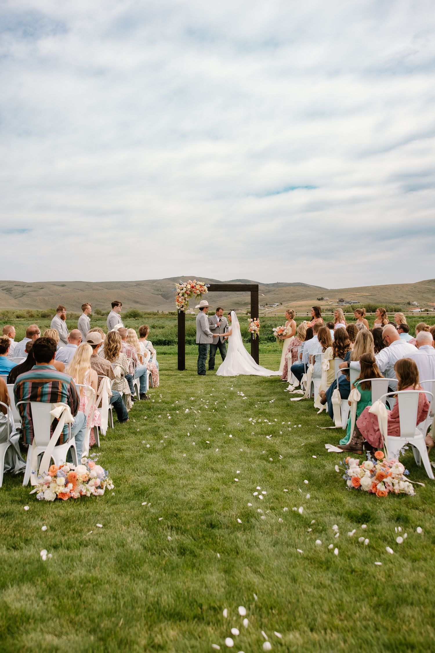 Outdoor wedding ceremony on green lawn with guests seated in white chairs against scenic mountain backdrop.