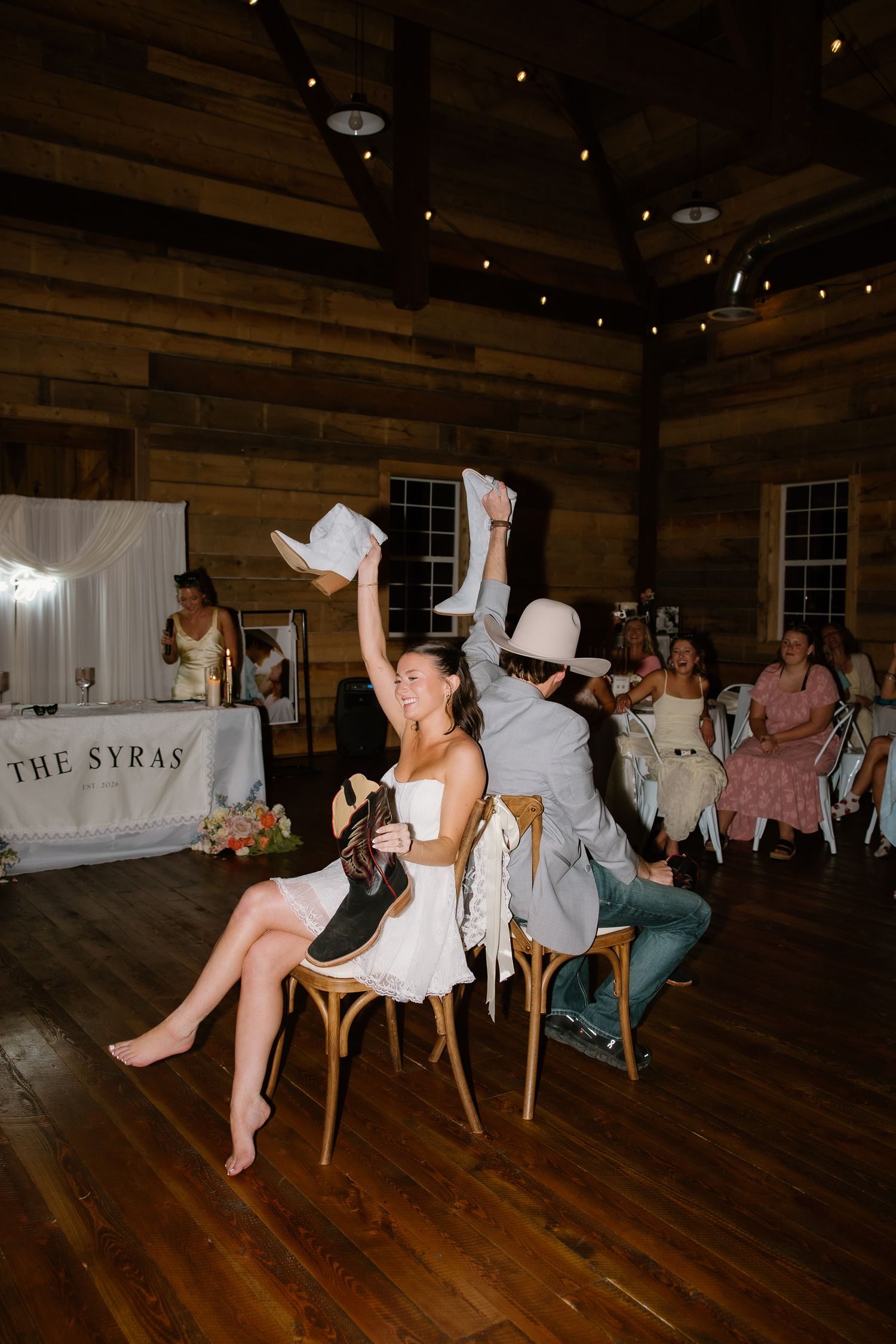 Bride and groom play the shoe game at a rustic barn wedding reception with guests watching.