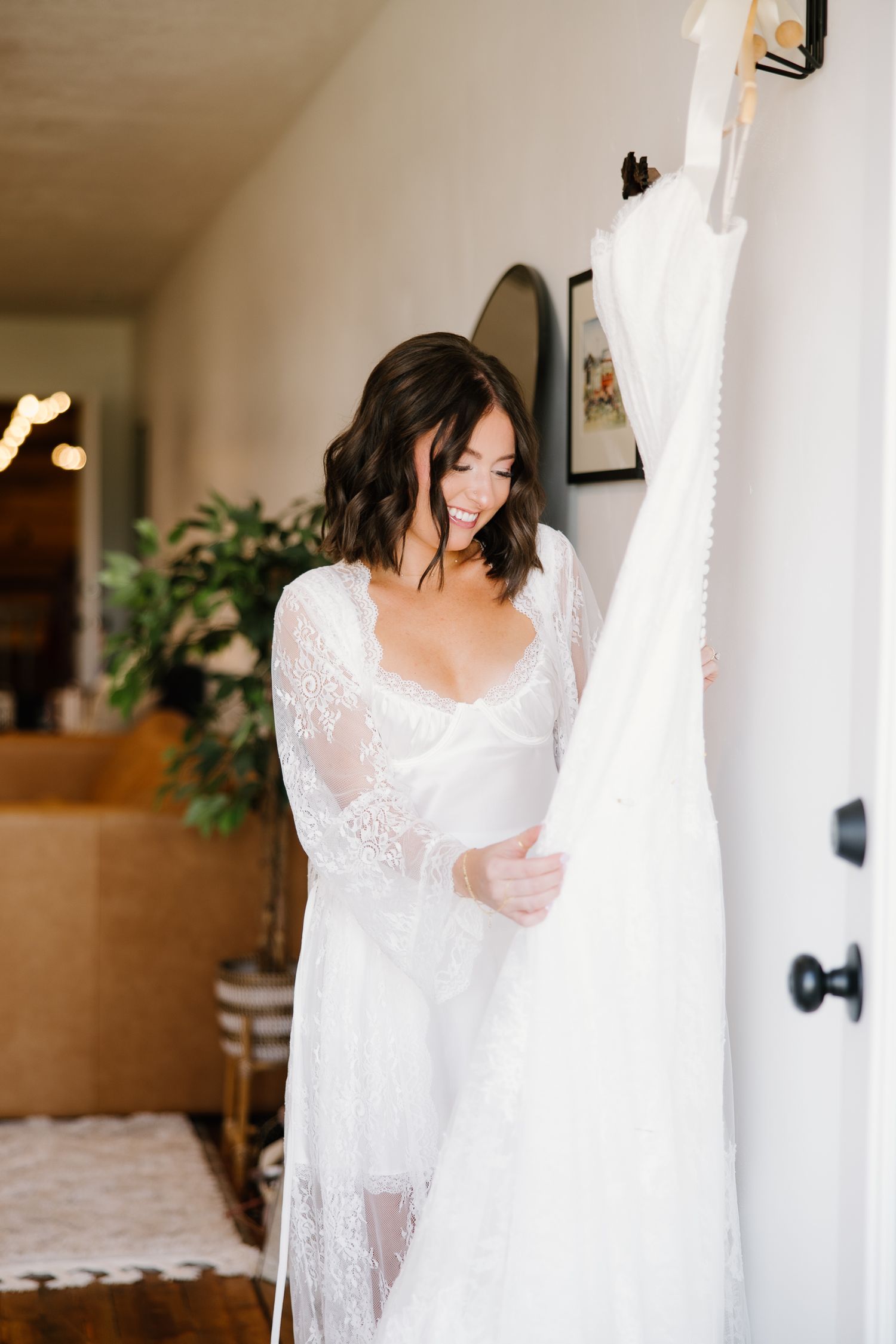 Bride in lace robe smiling at her wedding dress hanging on the wall before getting ready.