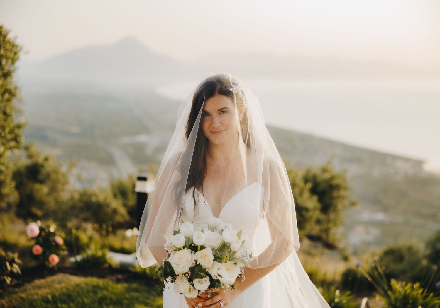 Bride in white wedding dress and veil holding white bouquet with scenic mountain and ocean backdrop.