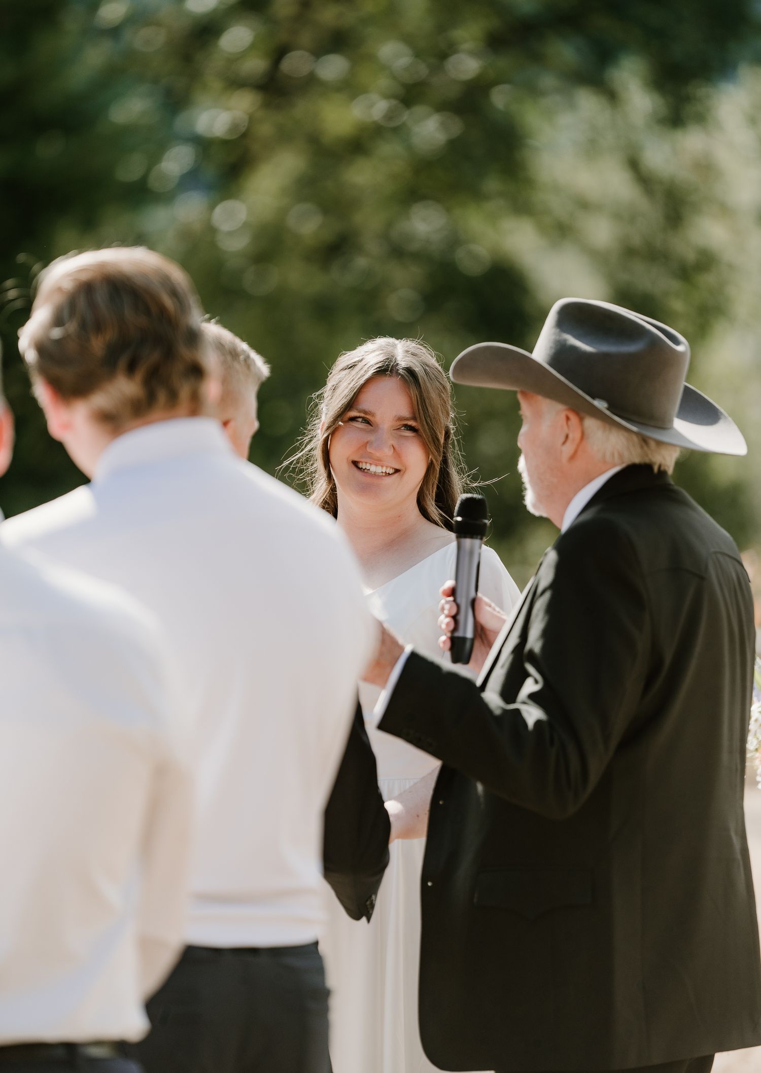 Bride smiles during outdoor wedding ceremony as officiant in black cowboy hat reads from notes.