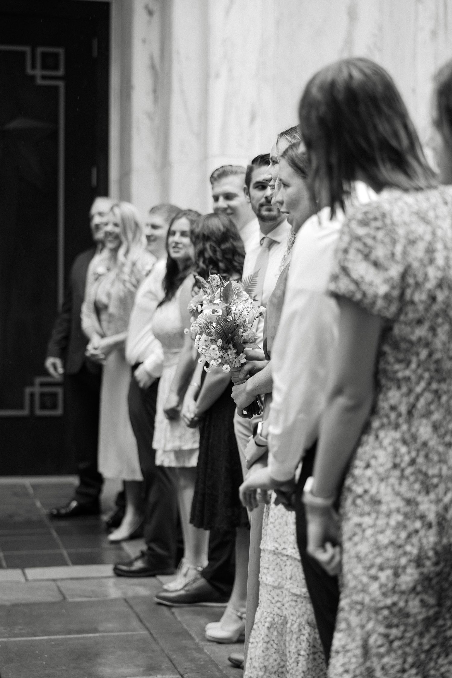 Black and white photo of a wedding party lineup outdoors, bride holding bouquet, guests in formal attire.