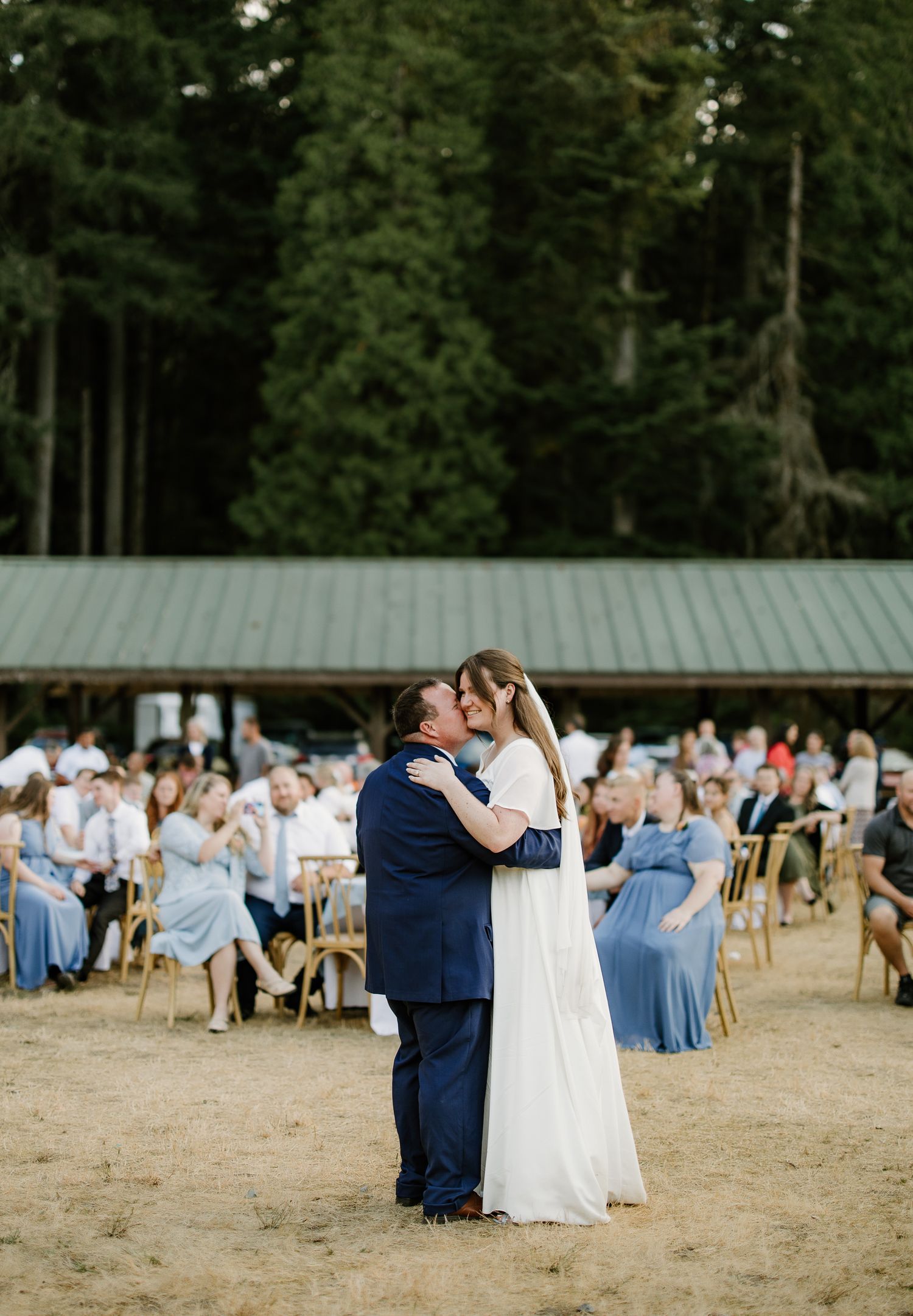 Bride and groom share first kiss at outdoor wedding ceremony with guests seated among tall trees.