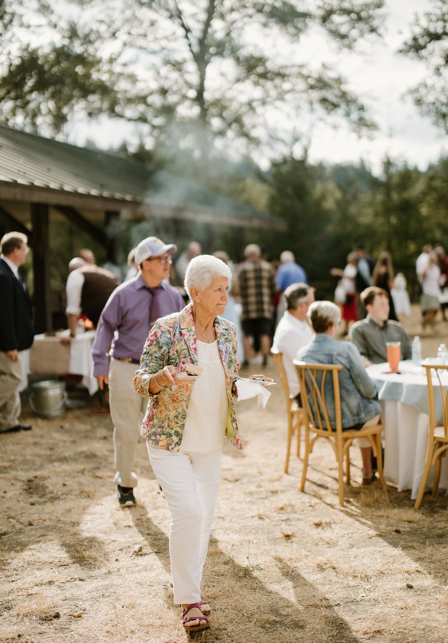Elderly woman in floral jacket and white pants walks at an outdoor rustic wedding reception with guests dining.