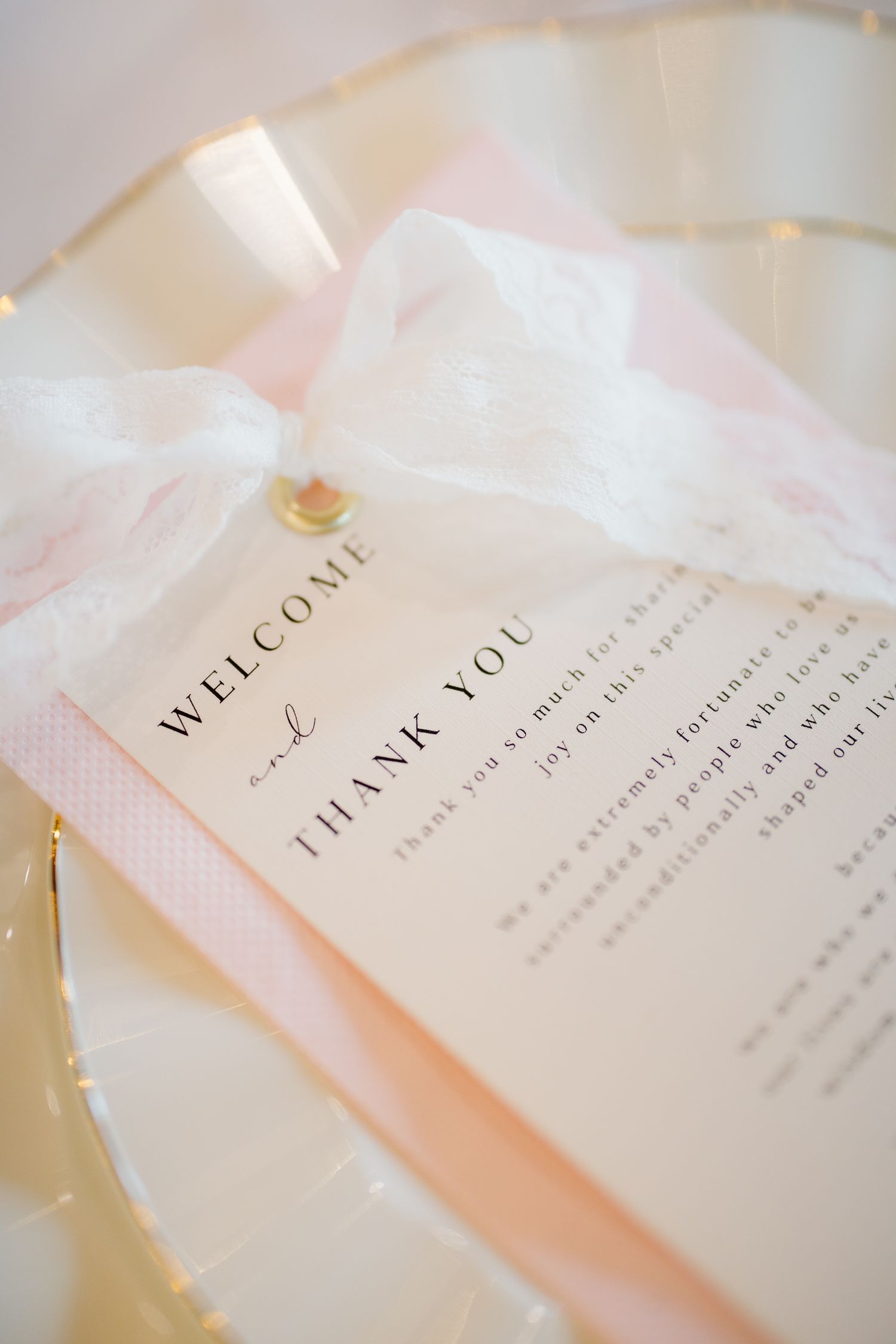 Elegant wedding welcome and thank you card on a gold plate with soft pink accents and delicate white ribbon.