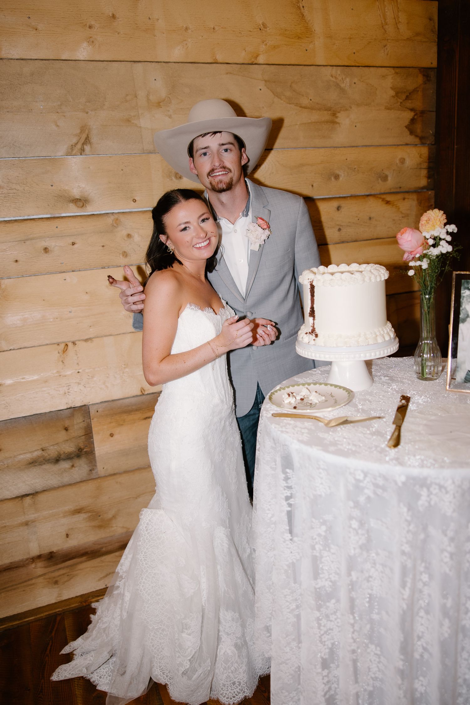 Smiling newlyweds cut white wedding cake together in rustic barn venue groom wearing cowboy hat.