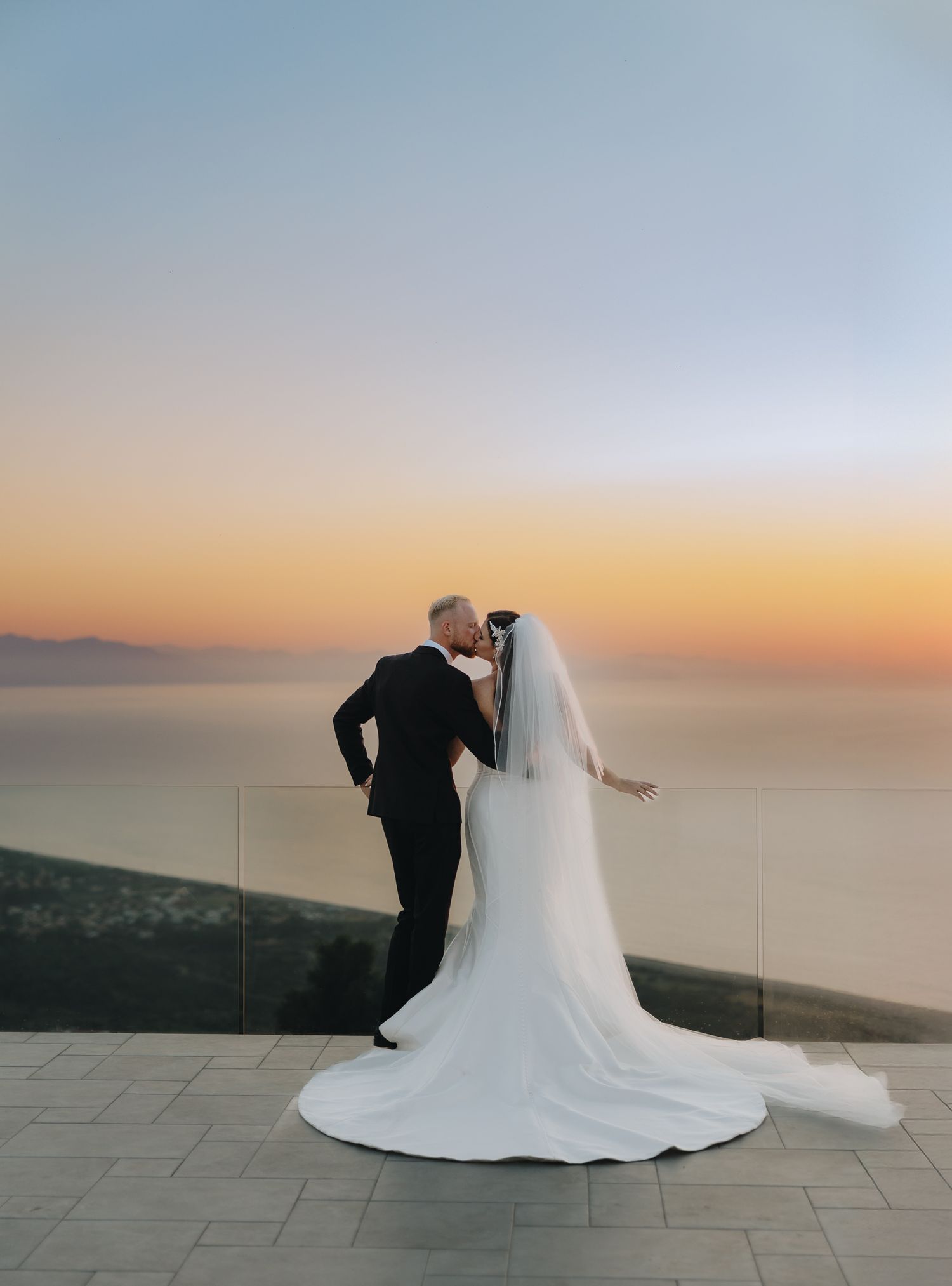 Bride and groom share a romantic kiss at sunset overlooking the ocean, with her elegant white gown and veil trailing.
