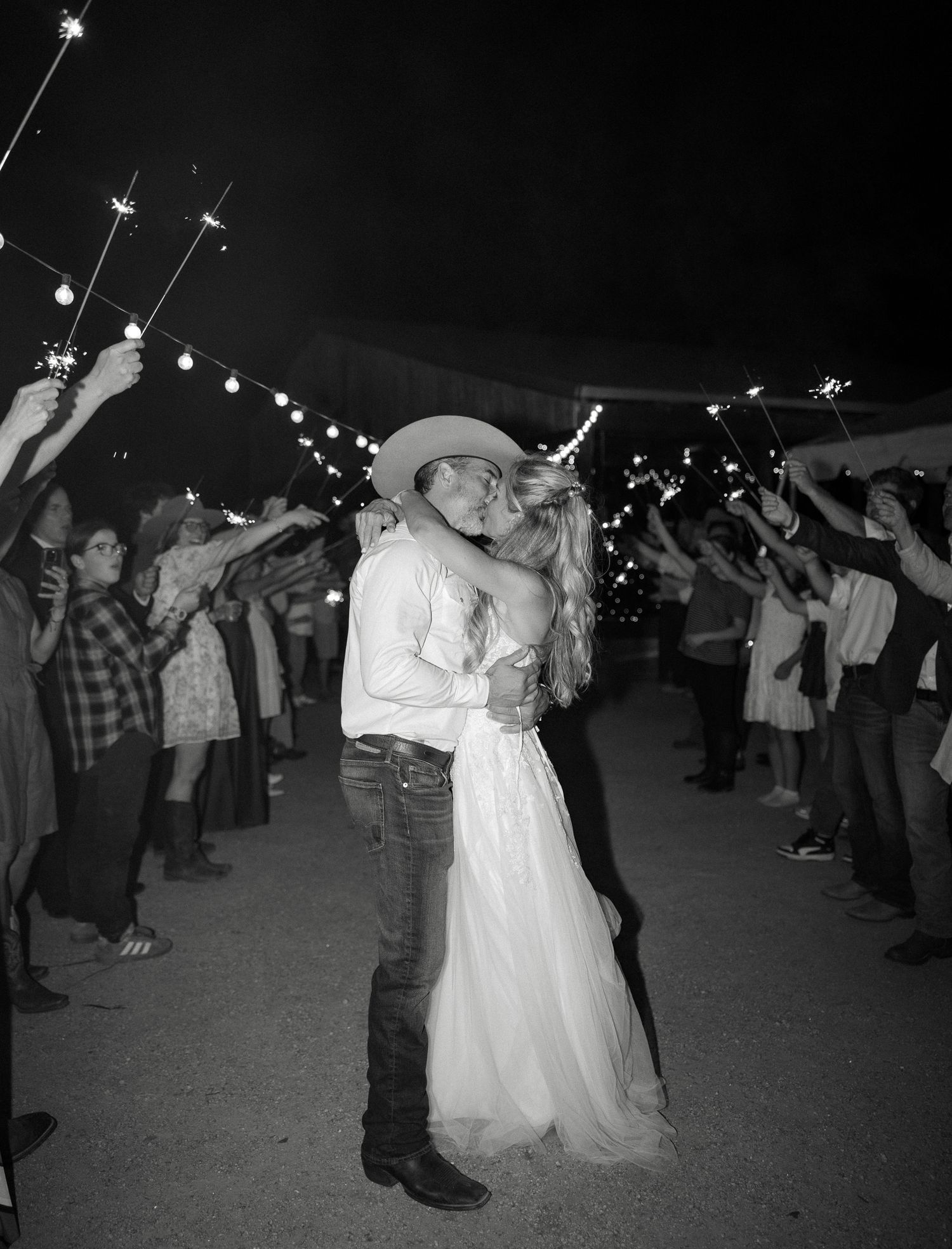 Black and white photo of bride and groom sharing romantic kiss during sparkler exit at outdoor evening wedding.