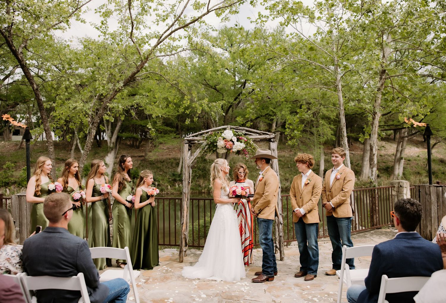 Outdoor rustic wedding ceremony with bride, groom, and wedding party under floral arch.