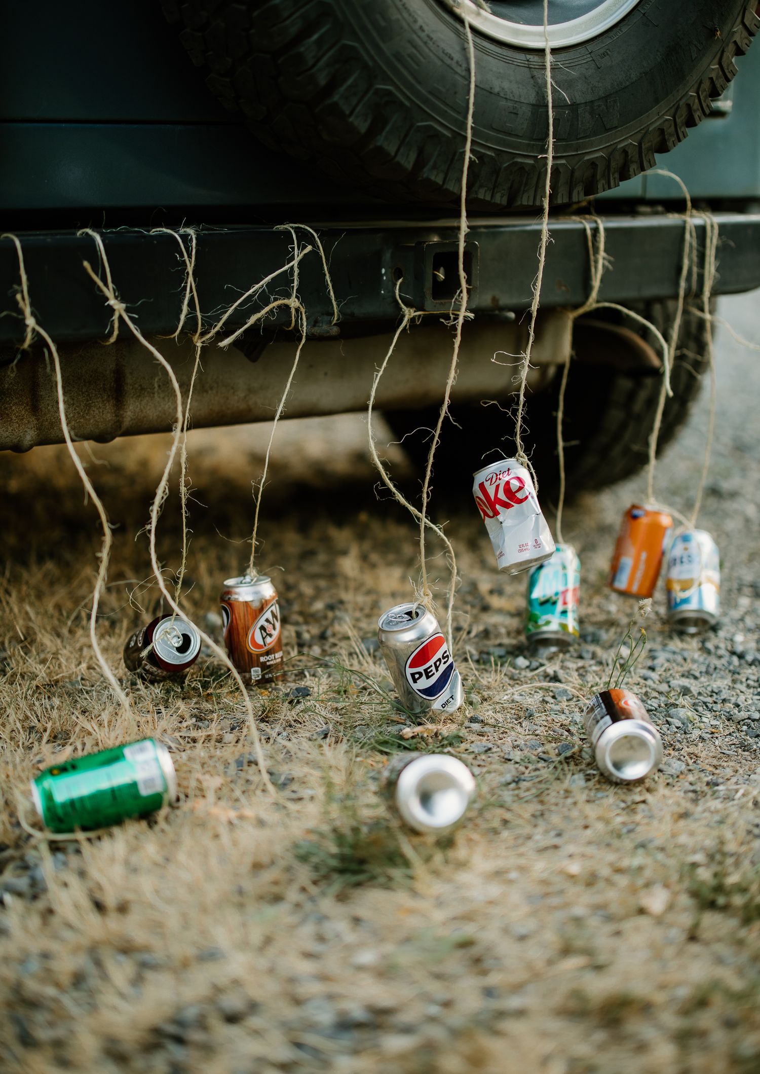 Soda cans tied with twine hanging from a car bumper as a wedding decoration on a dirt road.