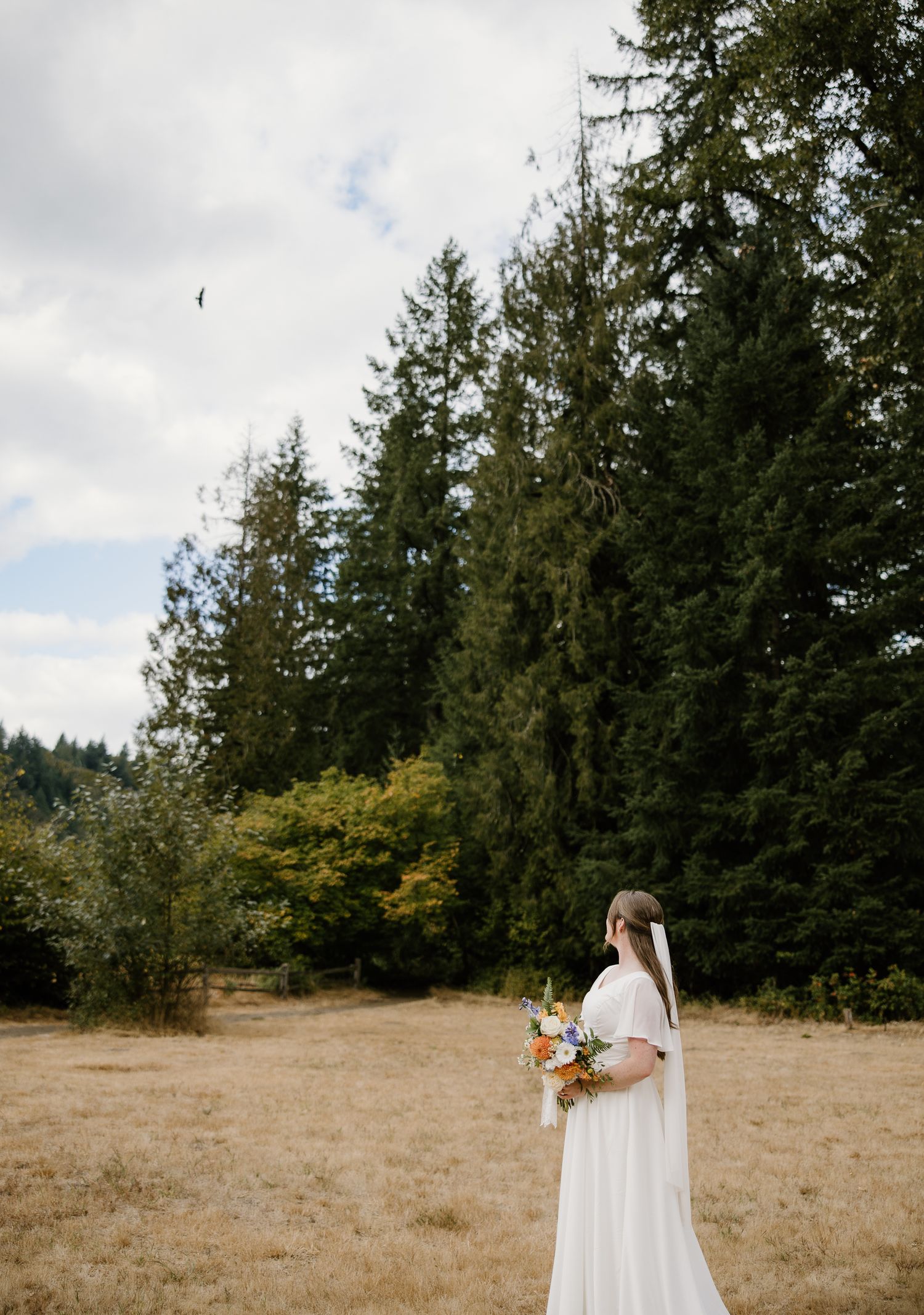 Bride in white gown holds colorful bouquet while standing in a dry meadow with evergreen trees.