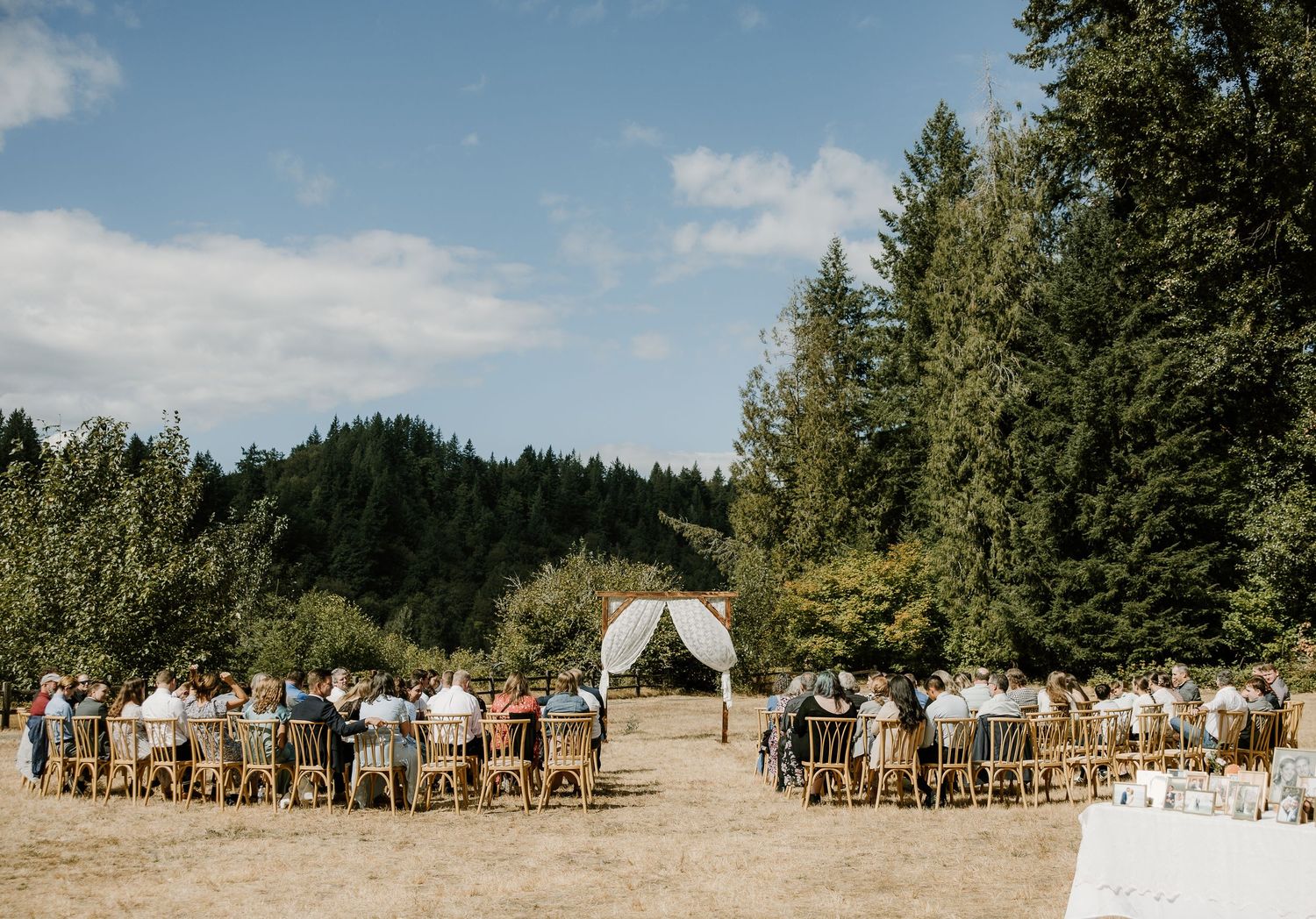 Outdoor wedding ceremony with guests seated on wooden chairs facing a floral arch in a sunny meadow.
