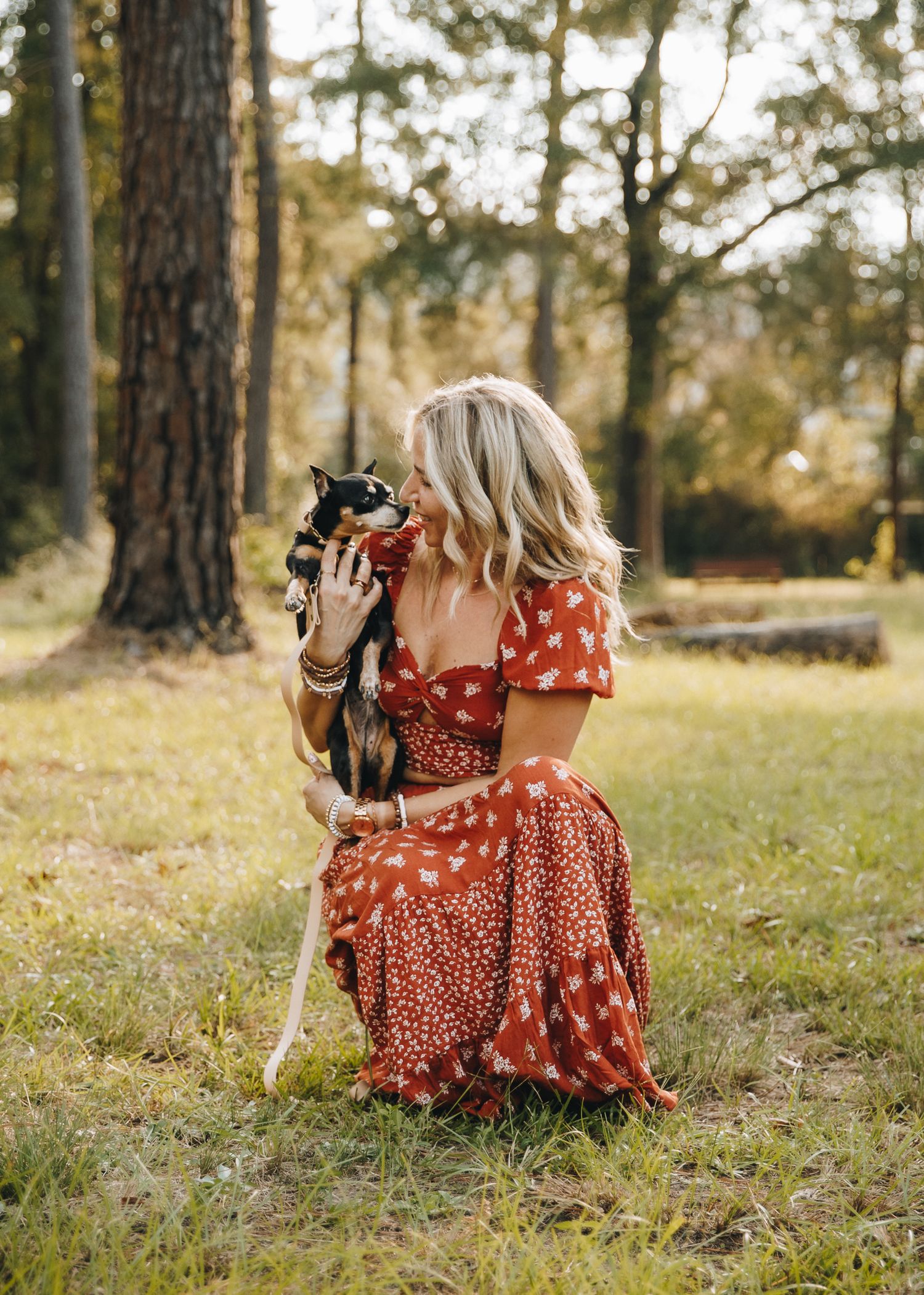Blonde woman in red floral dress sitting in a sunlit forest holding a small black and white cat.