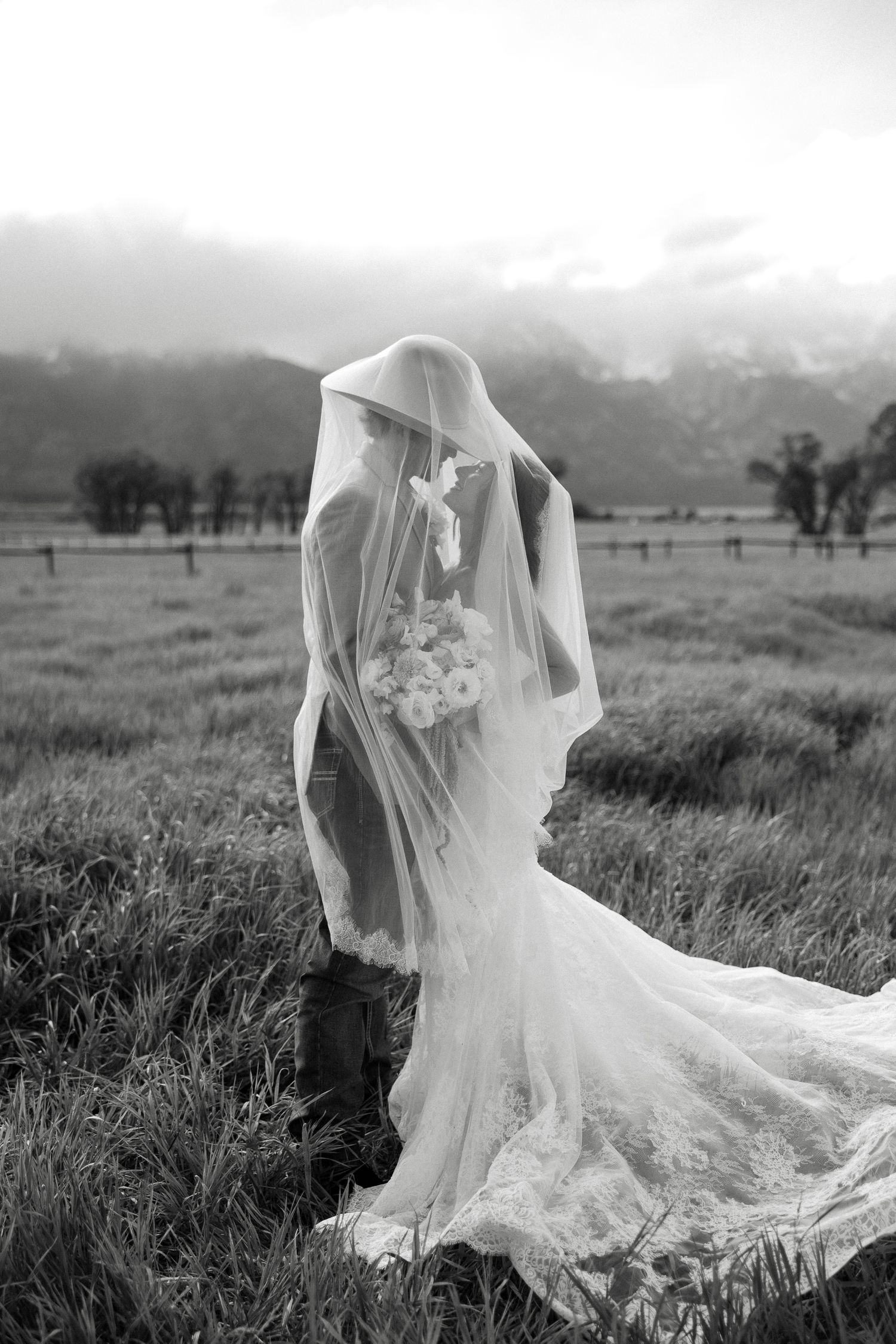 Black and white photo of bride and groom under veil in misty mountain meadow.