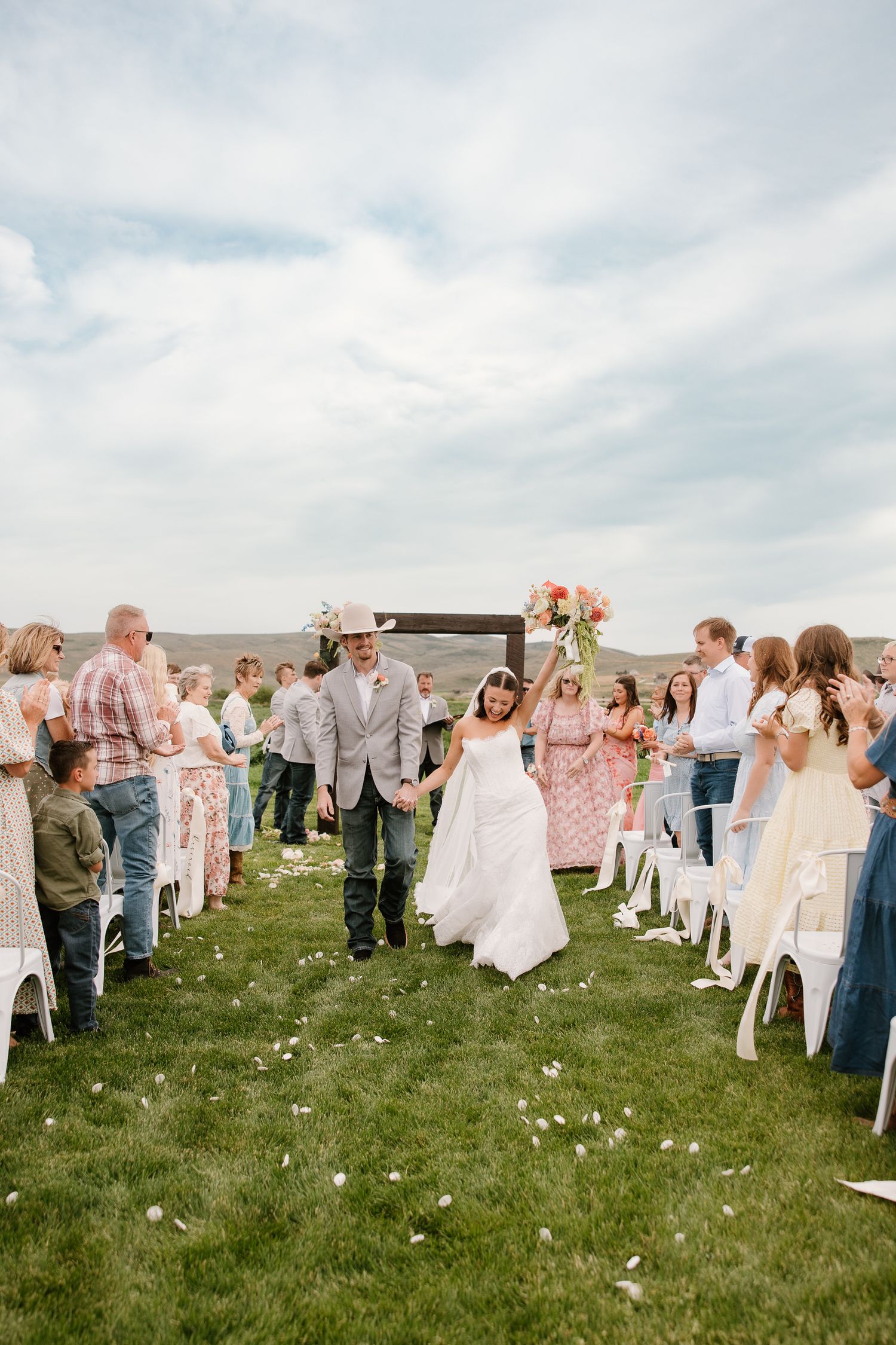Newlyweds walk back down the aisle through cheering guests after an outdoor wedding ceremony on a green lawn.