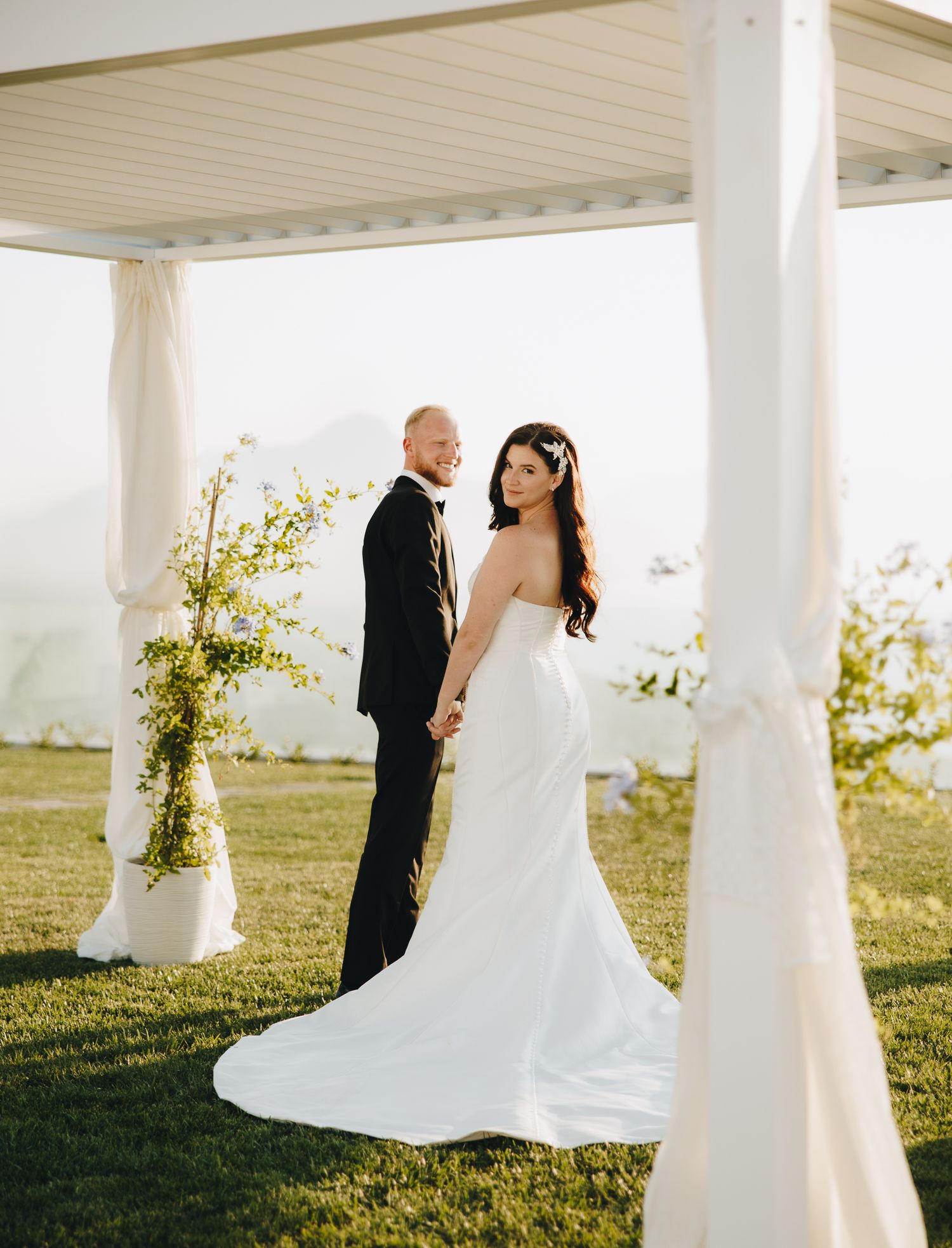 Bride and groom stand elegantly under a white draped pergola on green lawn, posing for wedding portrait.
