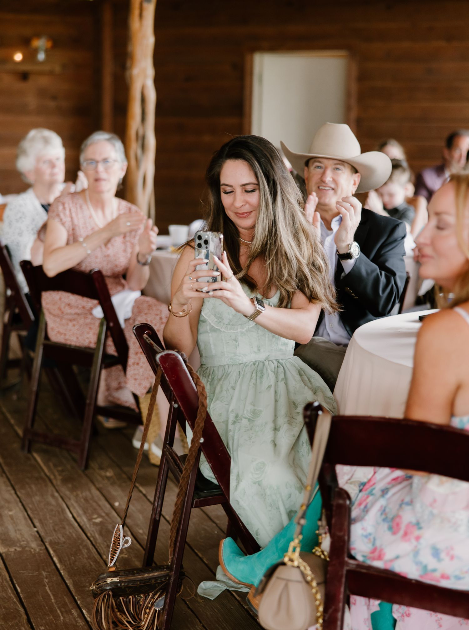 Wedding reception guests at rustic wooden venue, woman photographing with smartphone.