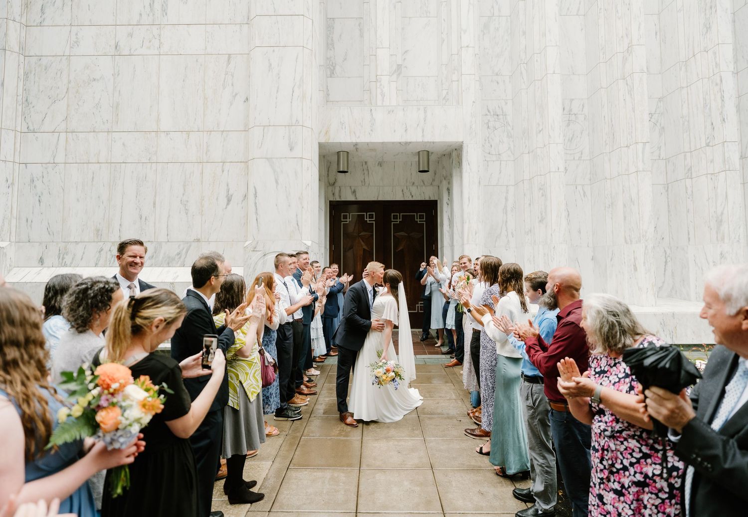 Newlyweds kiss while guests line up to celebrate outside a grand marble building during a wedding ceremony.