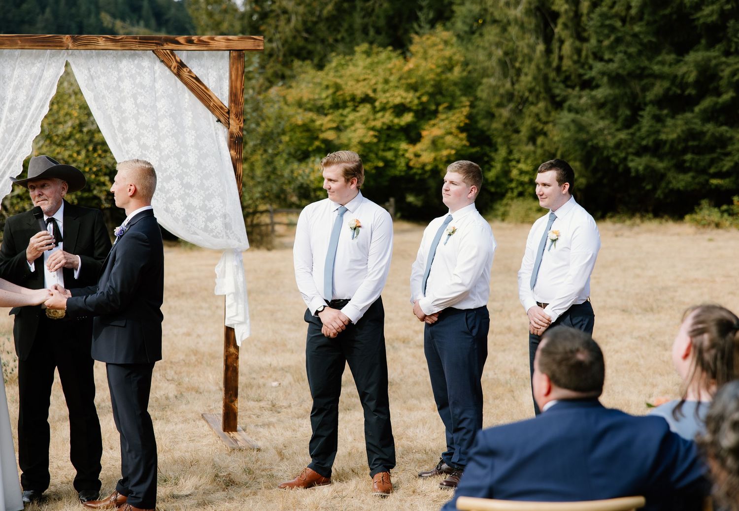 Groomsmen in white shirts and blue ties stand at outdoor wedding ceremony altar with rustic wooden arch.