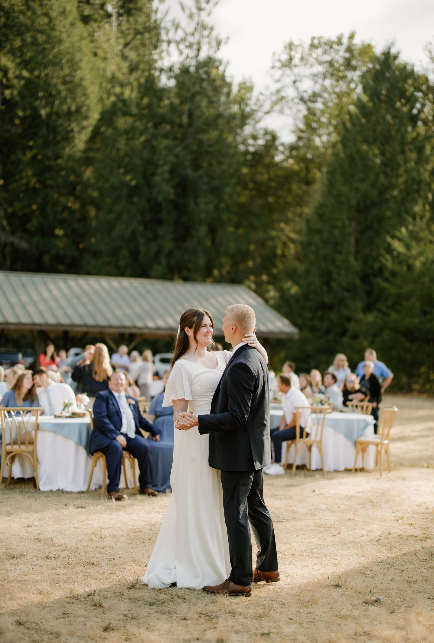 Bride and groom share first dance outdoors at elegant wedding reception with guests seated at white-draped tables amid tall trees.