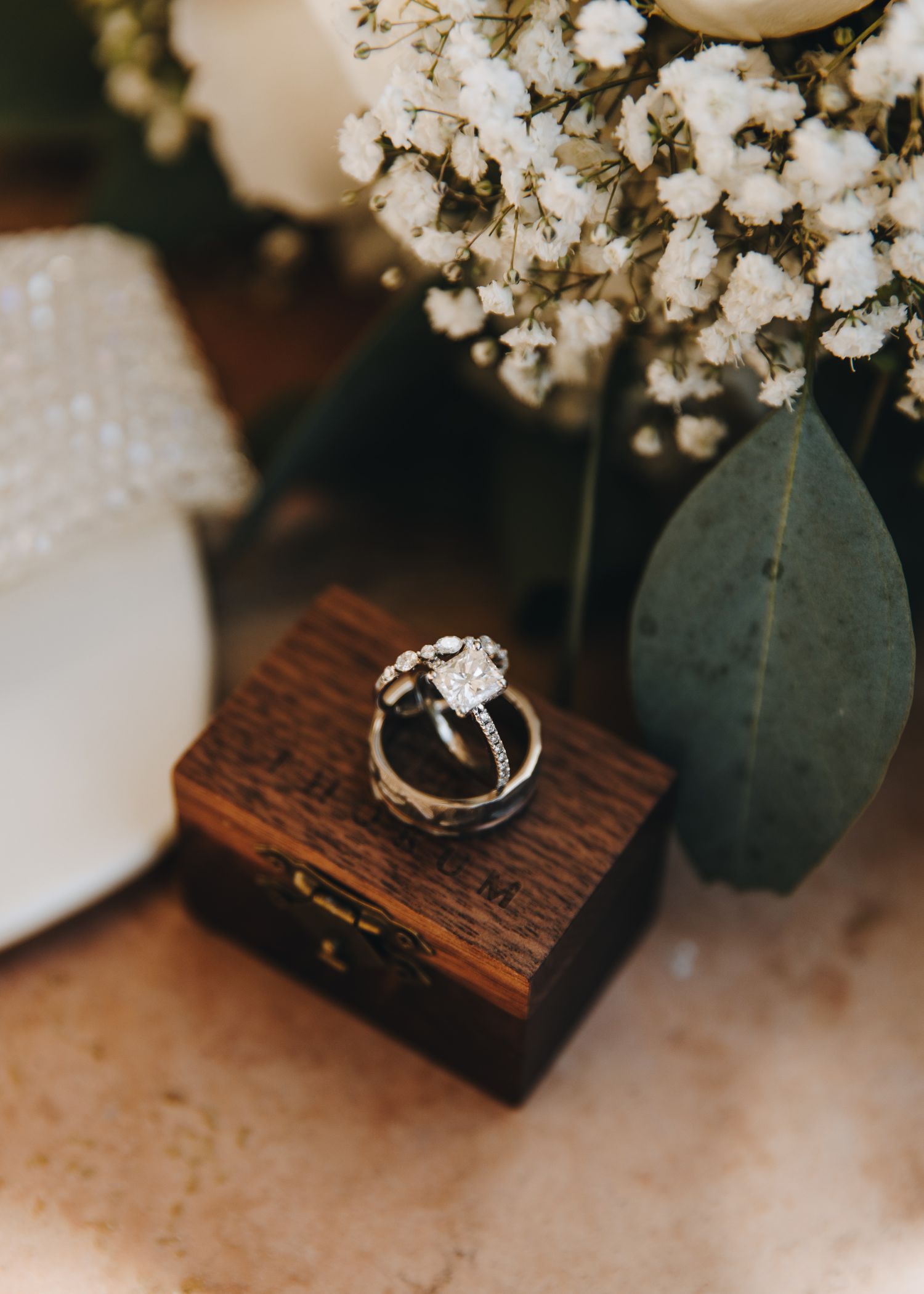 Diamond engagement ring and wedding bands displayed on rustic wooden ring box with flowers.
