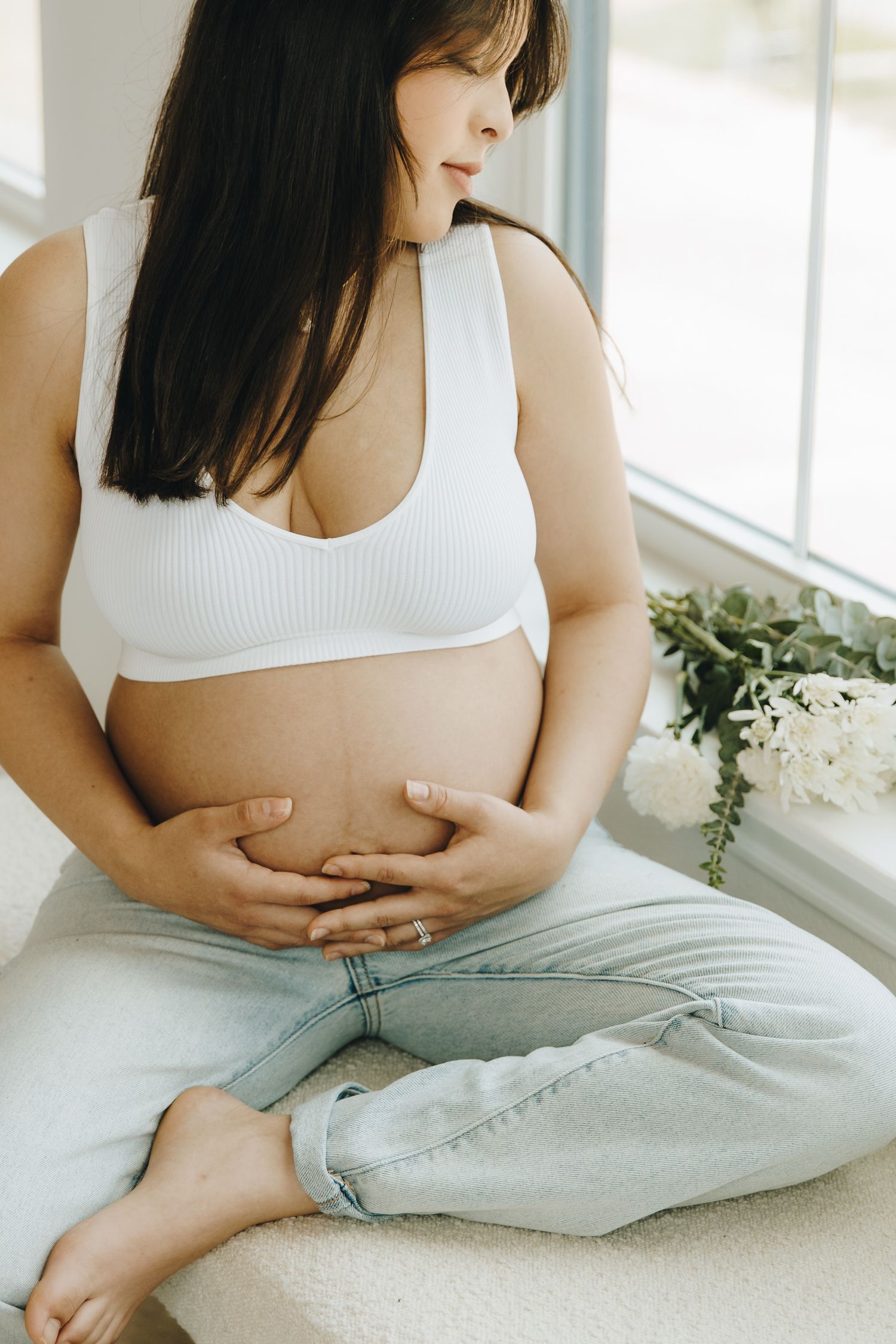 Pregnant woman in white crop top and jeans sitting cross-legged, cradling her baby bump.