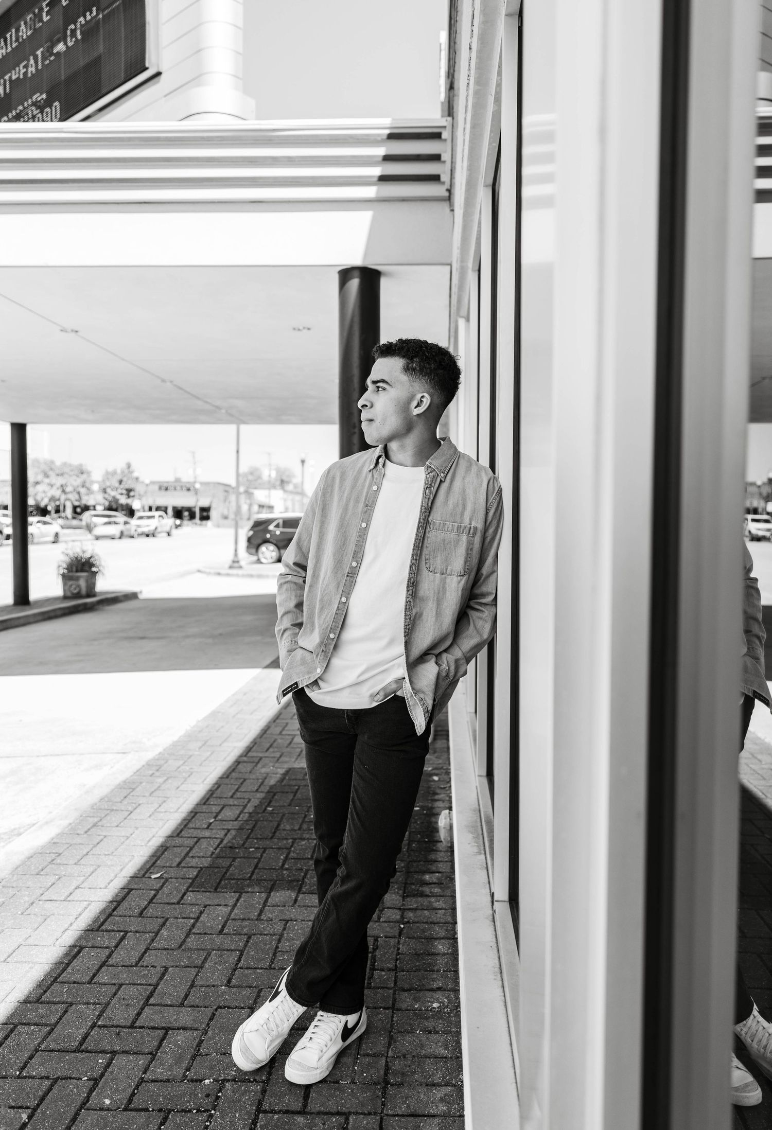 Black and white photo of young man leaning against building on urban sidewalk.
