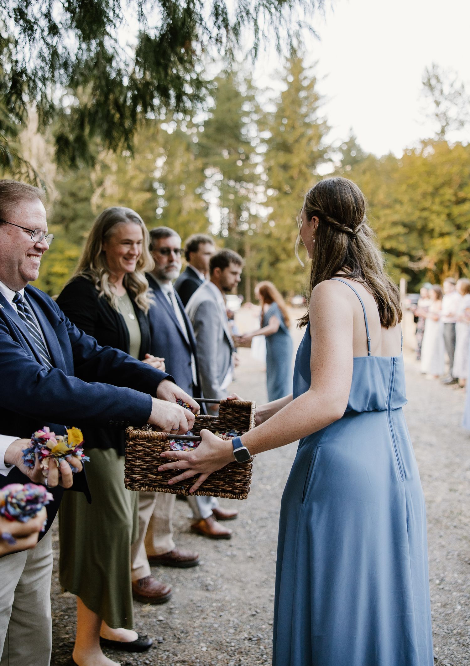 Bridesmaid in blue gown handing flower petals from a wicker basket to smiling wedding guests outdoors.