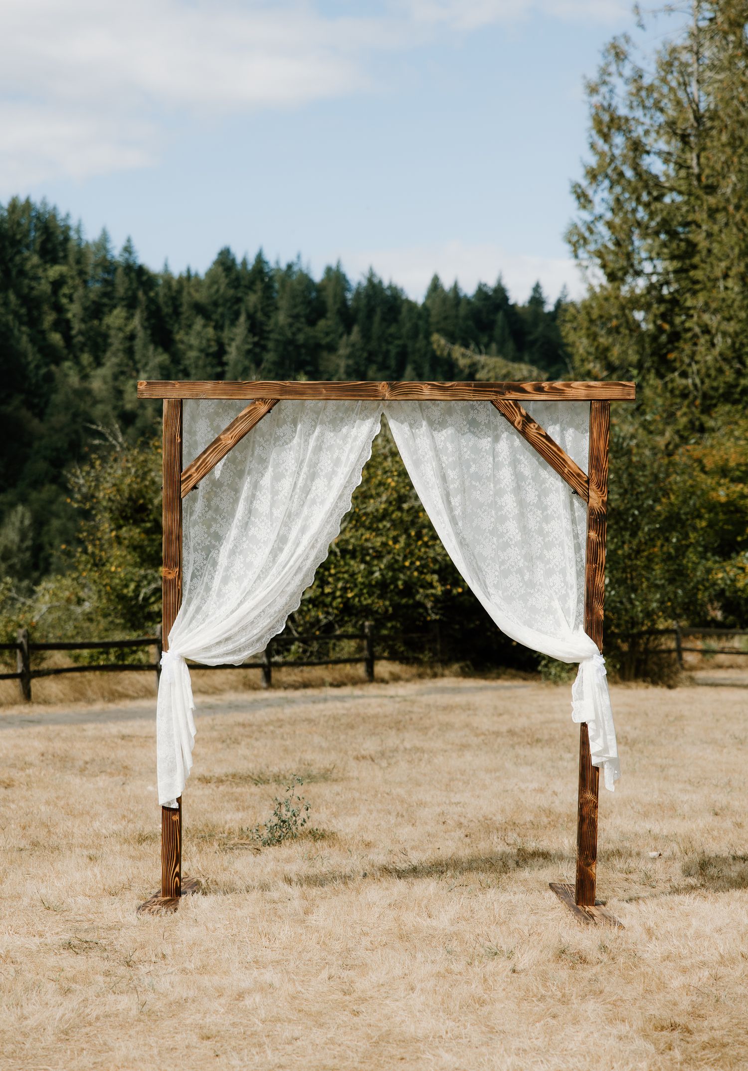 Rustic wooden wedding arch draped with white lace curtains stands in a golden meadow.