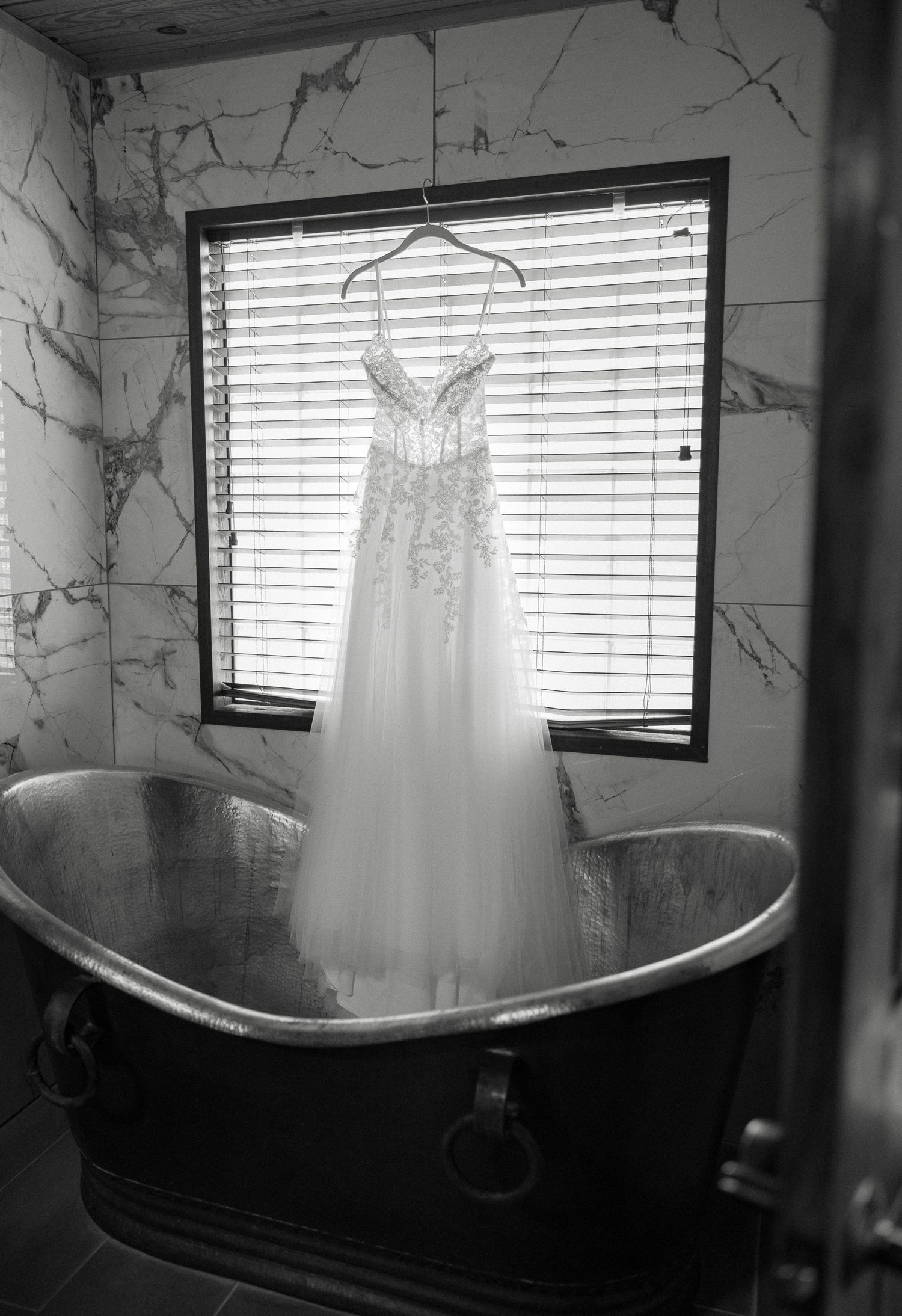 Black and white photo of an elegant lace wedding dress hanging over a vintage bathtub in a marble bathroom.