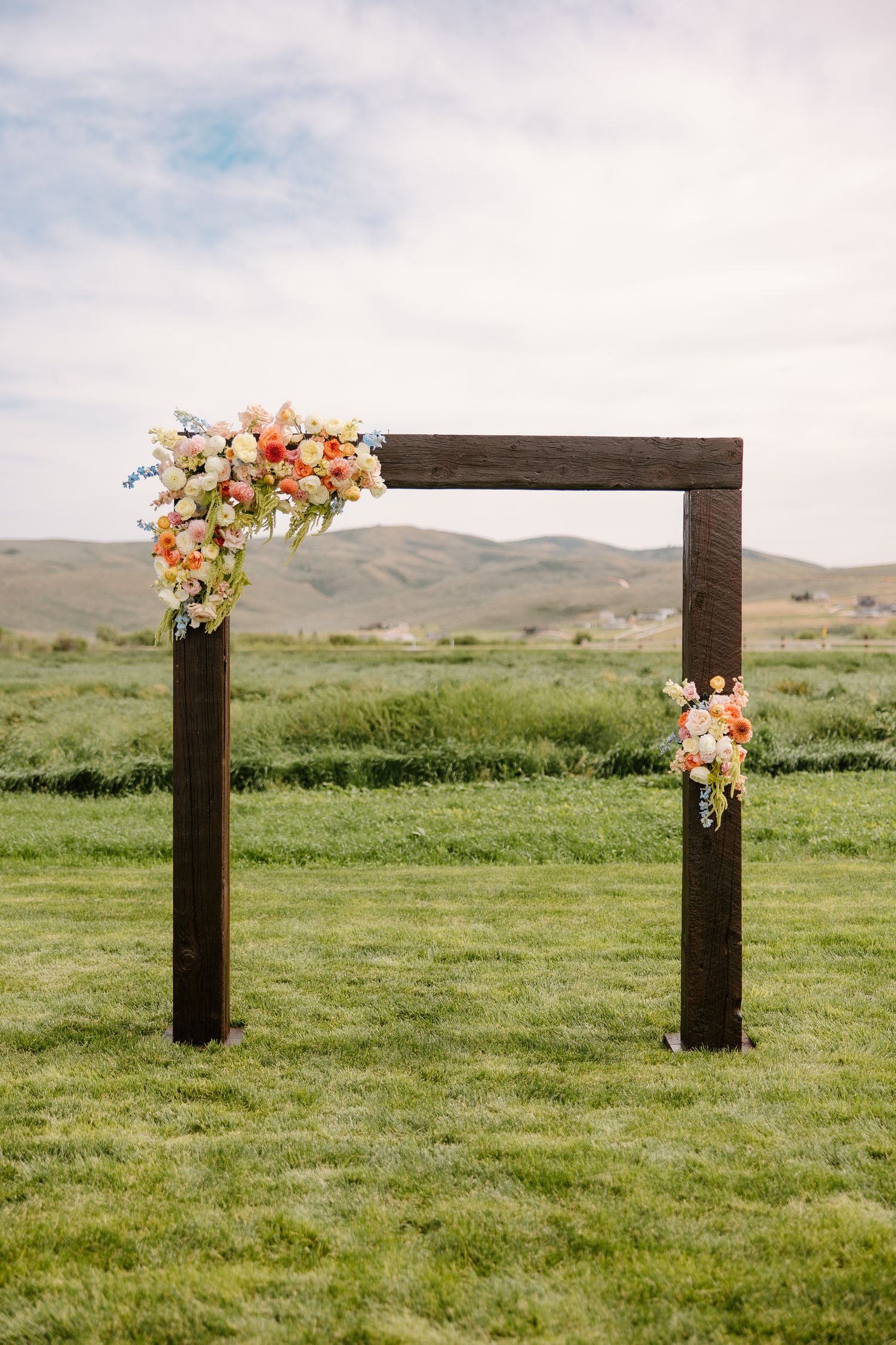 Rustic dark wood wedding arch adorned with peach and pink floral arrangements on a green lawn with rolling hills.