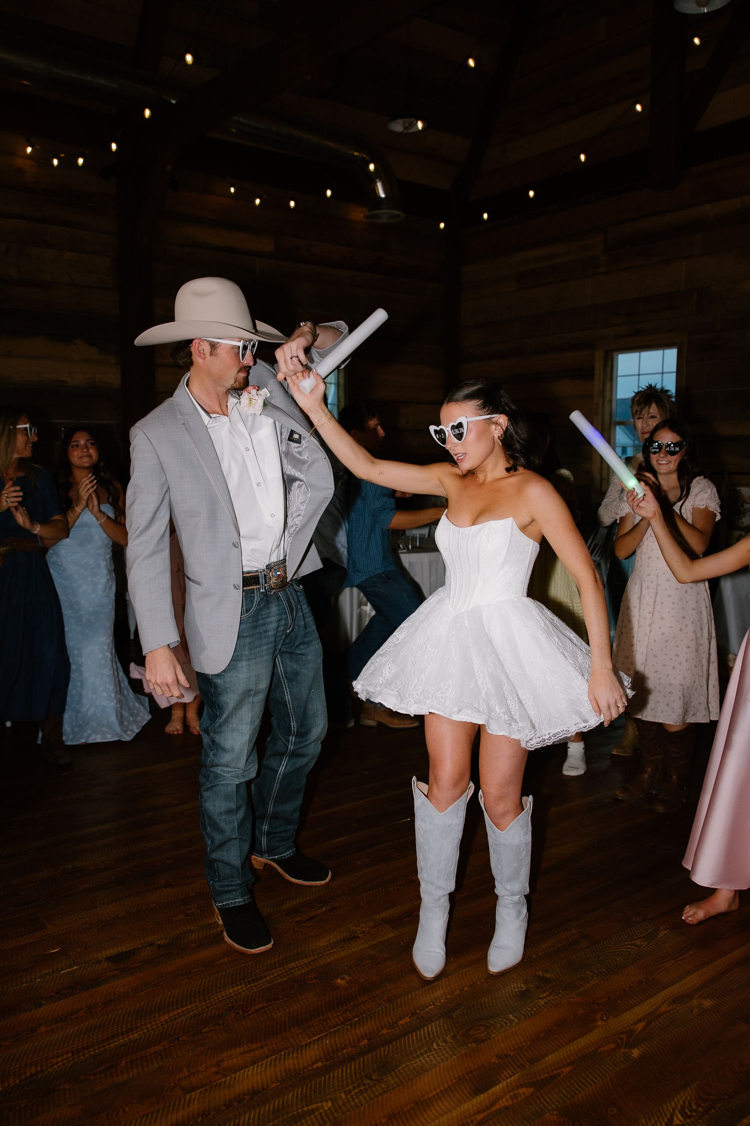 Bride in white mini dress and cowboy boots dances with groom in gray suit and cowboy hat at rustic barn wedding reception.