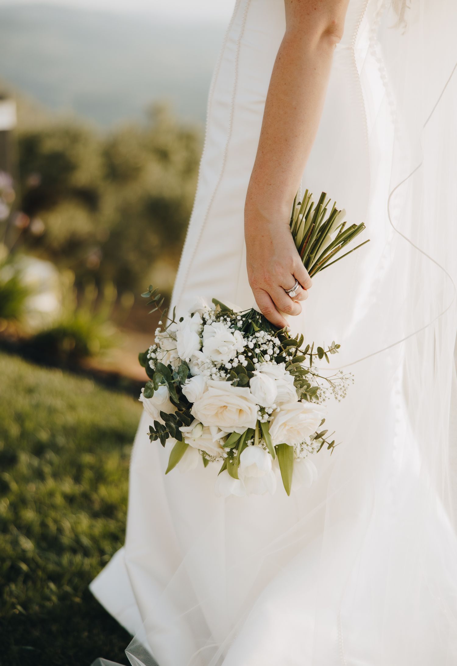 Bride in white wedding gown holding elegant bouquet of white roses and greenery outdoors.