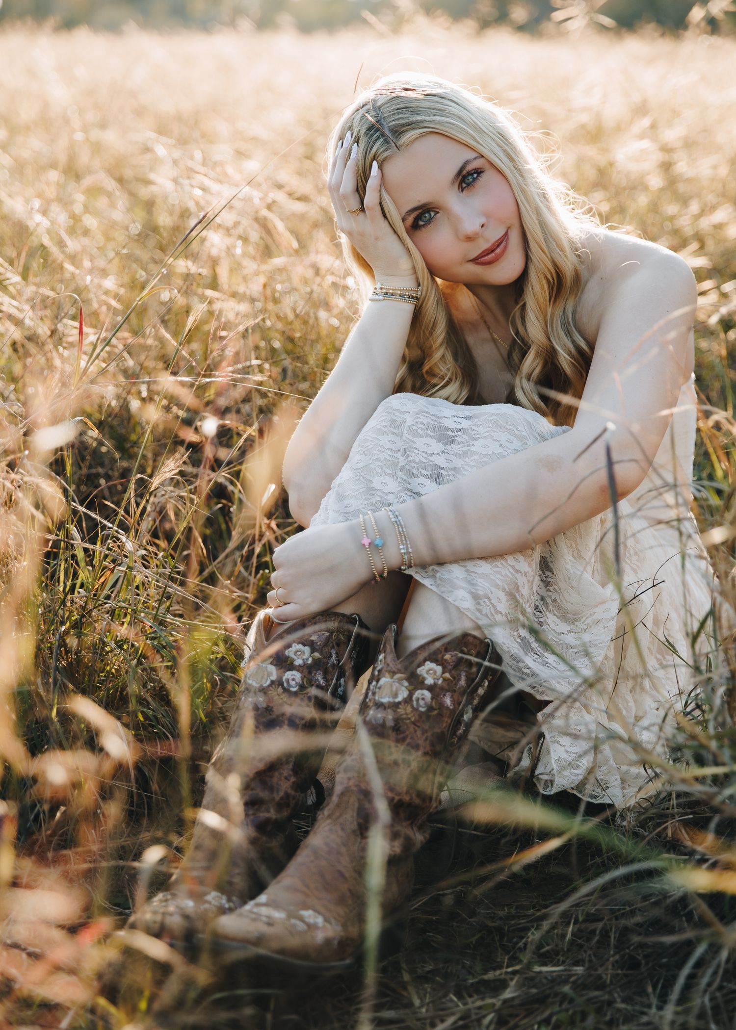 Blonde woman in white lace outfit and cowboy boots sitting in golden field at sunset.