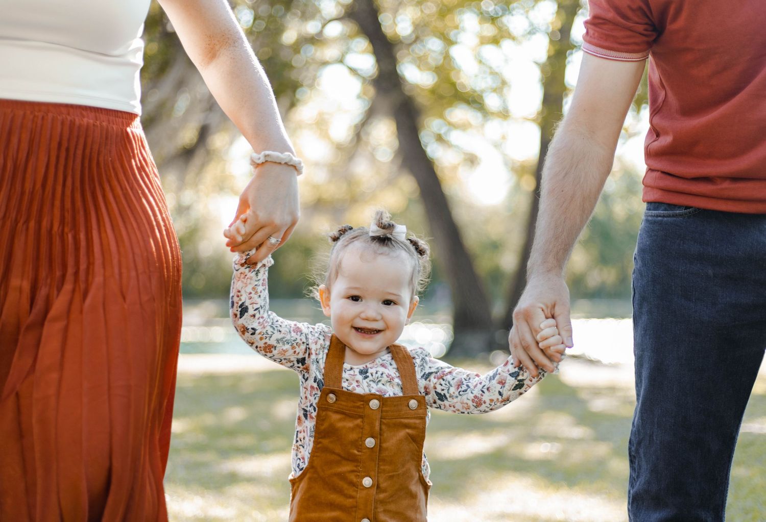 Smiling toddler girl in overalls holding hands with parents during a sunny outdoor family photo session.