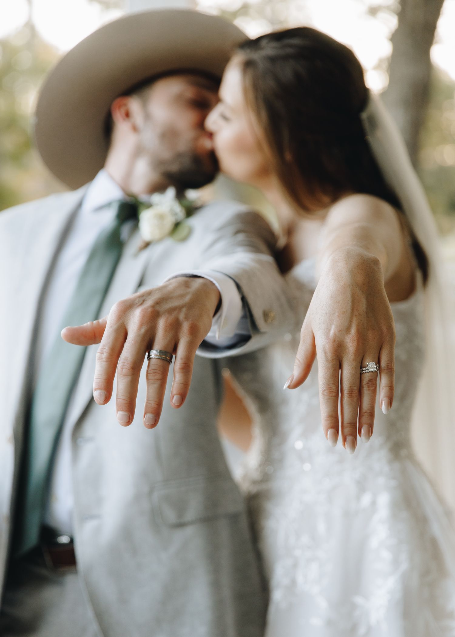 Bride and groom kissing while showing off wedding rings to camera at outdoor country wedding.