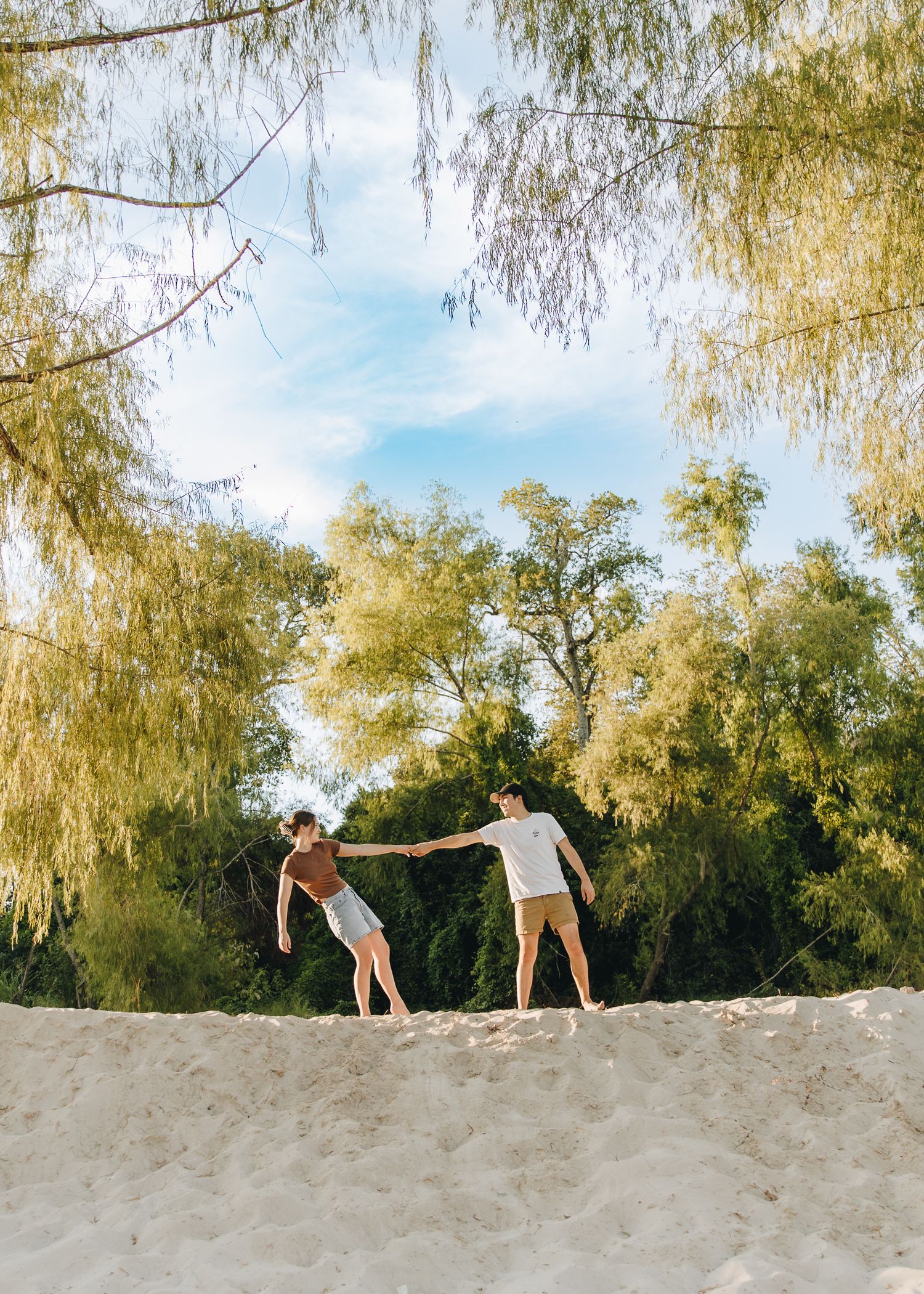 Couple holding hands while walking on a sandy beach framed by willow trees under a blue sky.