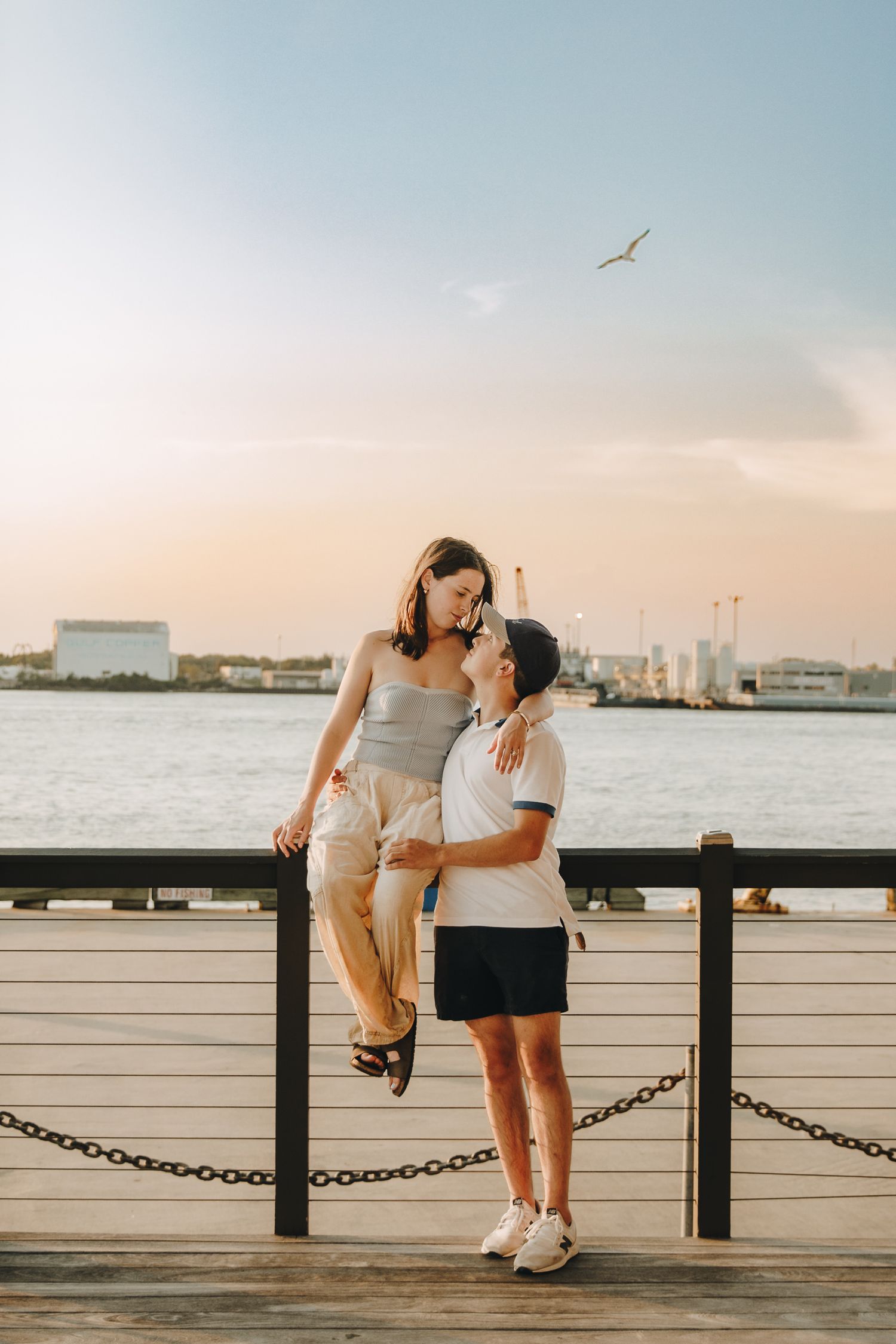 Couple sharing a tender moment on a waterfront pier at sunset, woman seated on railing.