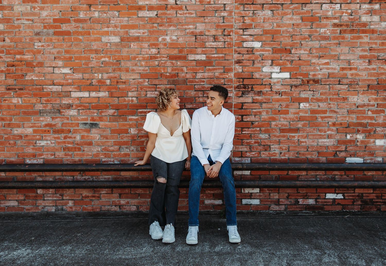Couple sitting on bench against red brick wall, smiling at each other.