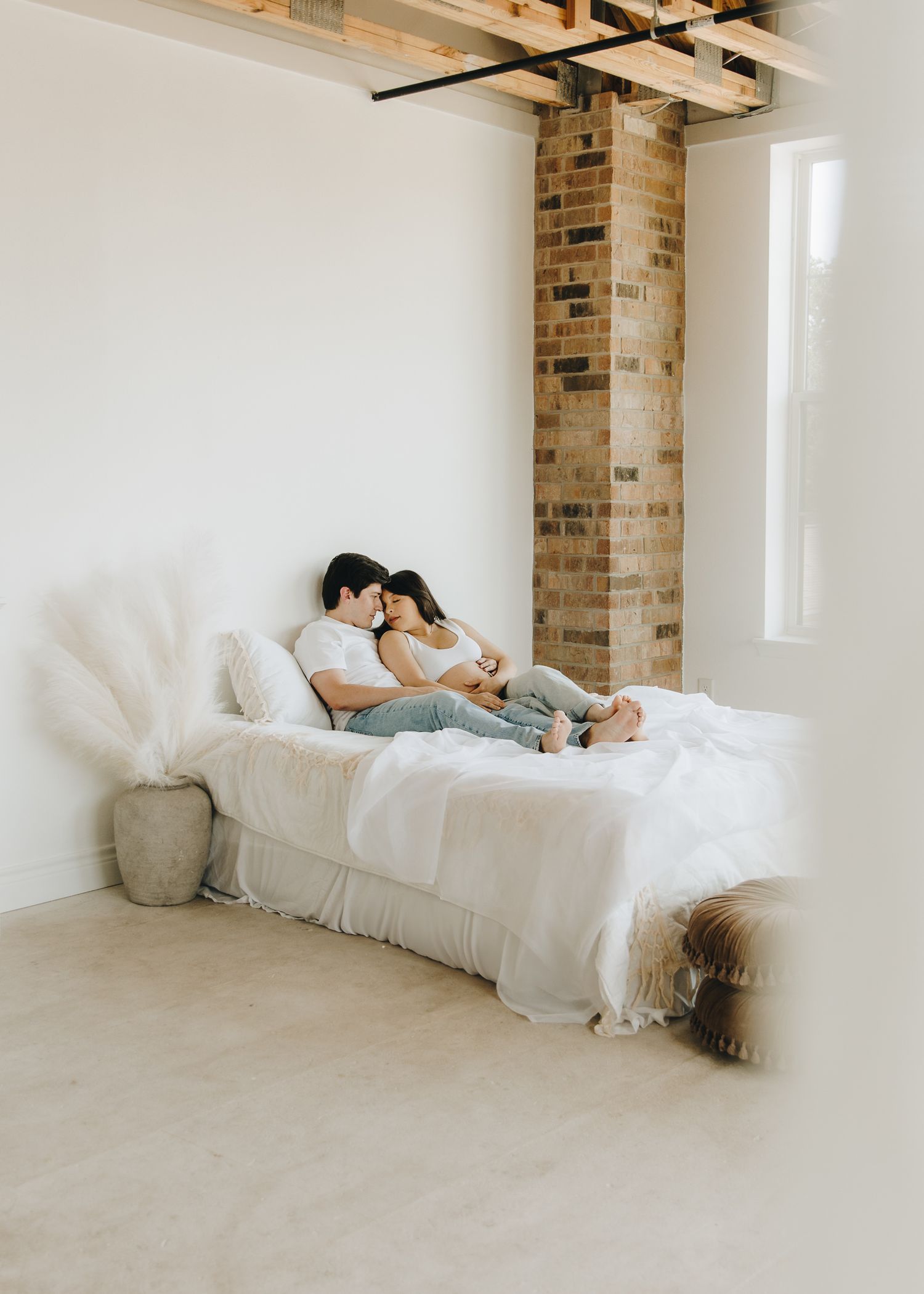 Expecting couple cuddling on a white bed in a bright loft with exposed brick pillars.