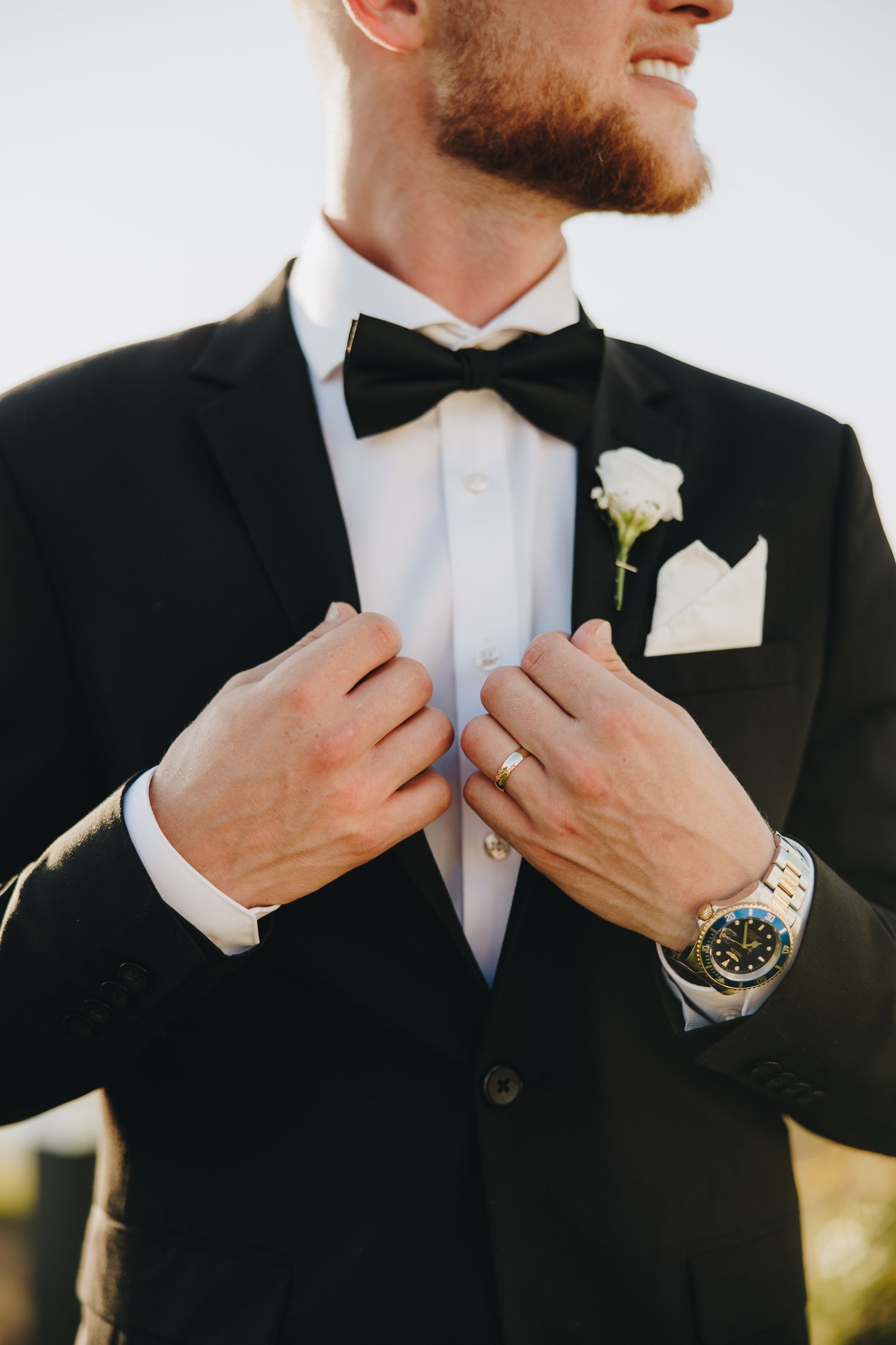 Smiling groom in black tuxedo adjusting lapels, wearing bow tie, boutonniere, wedding ring, and luxury watch.