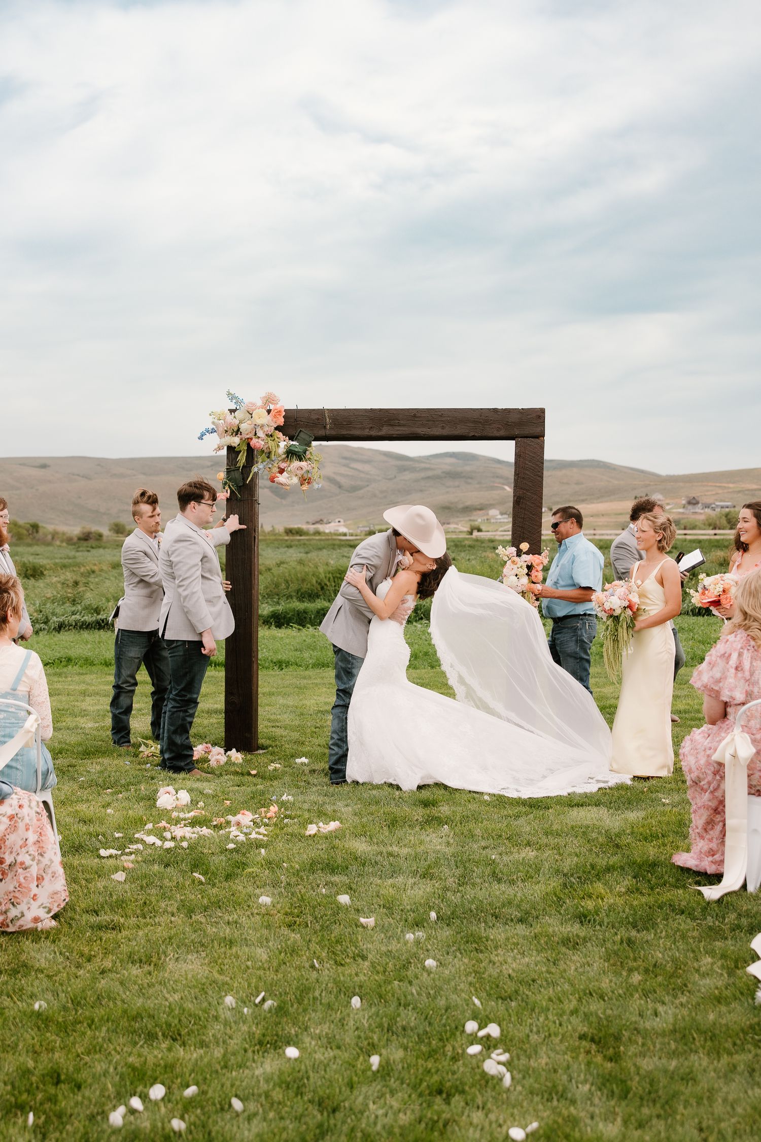 Bride and groom share first kiss under rustic wooden arch adorned with flowers at outdoor wedding ceremony.