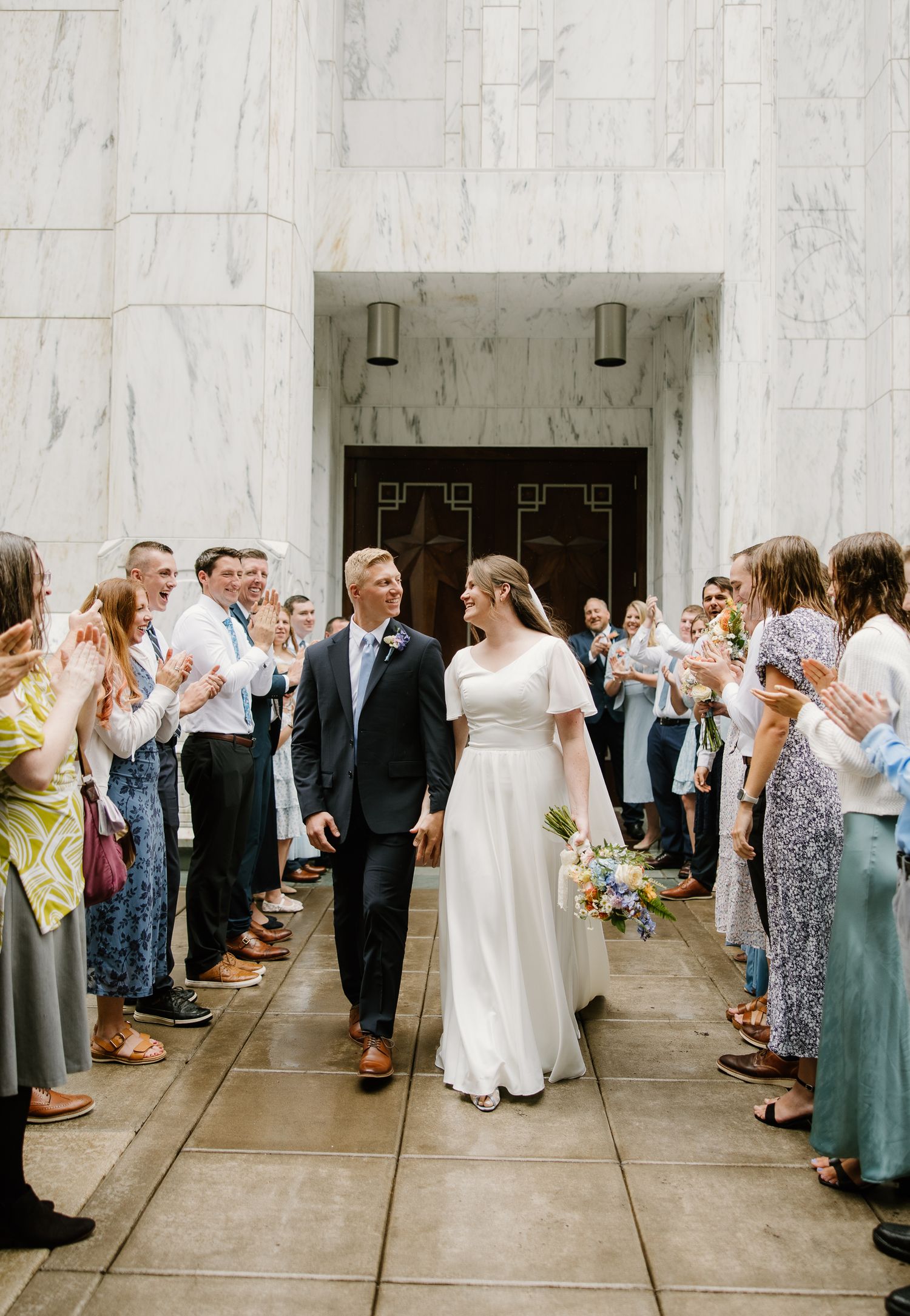 Bride and groom walk through cheering guests outside a marble building after their wedding ceremony.