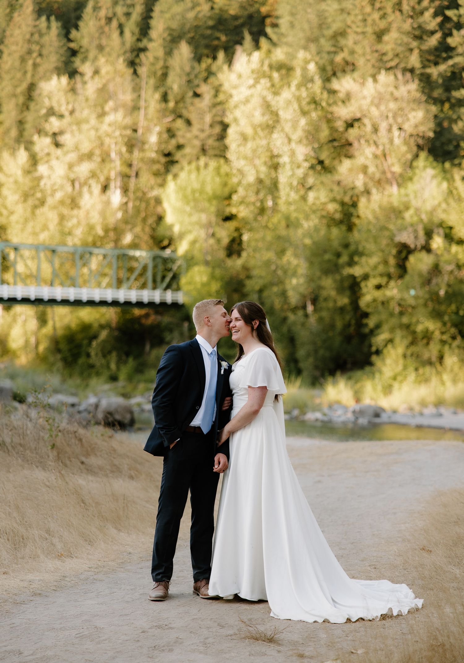 Bride and groom smile at each other on a sandy path with a green bridge and river in the background.