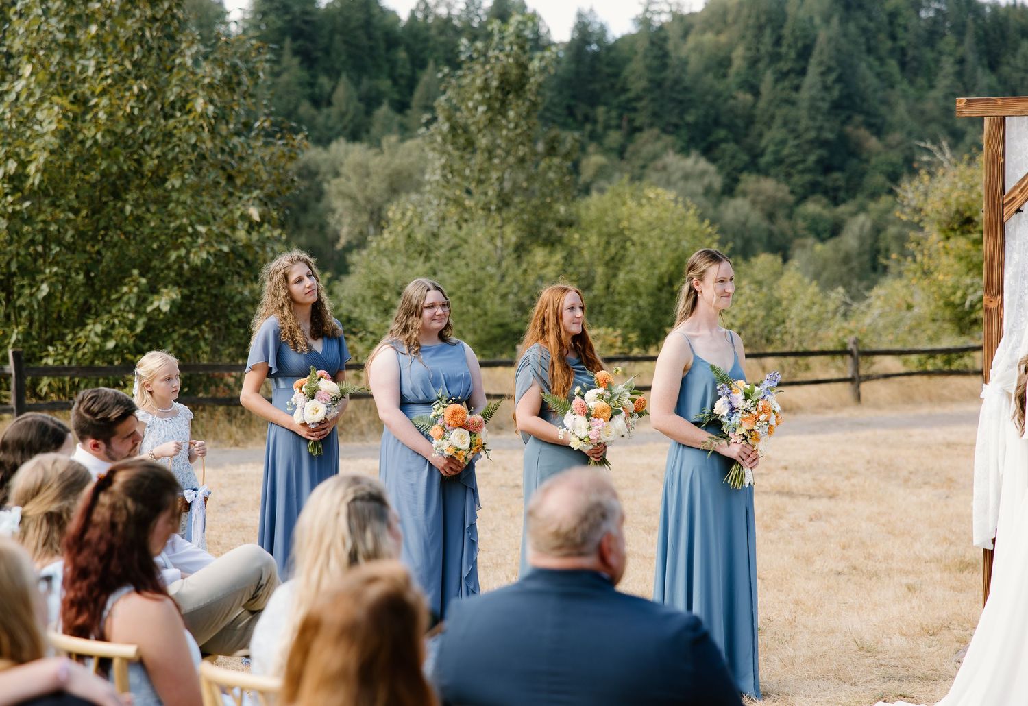 Four bridesmaids in blue dresses holding colorful bouquets stand at an outdoor wedding ceremony.
