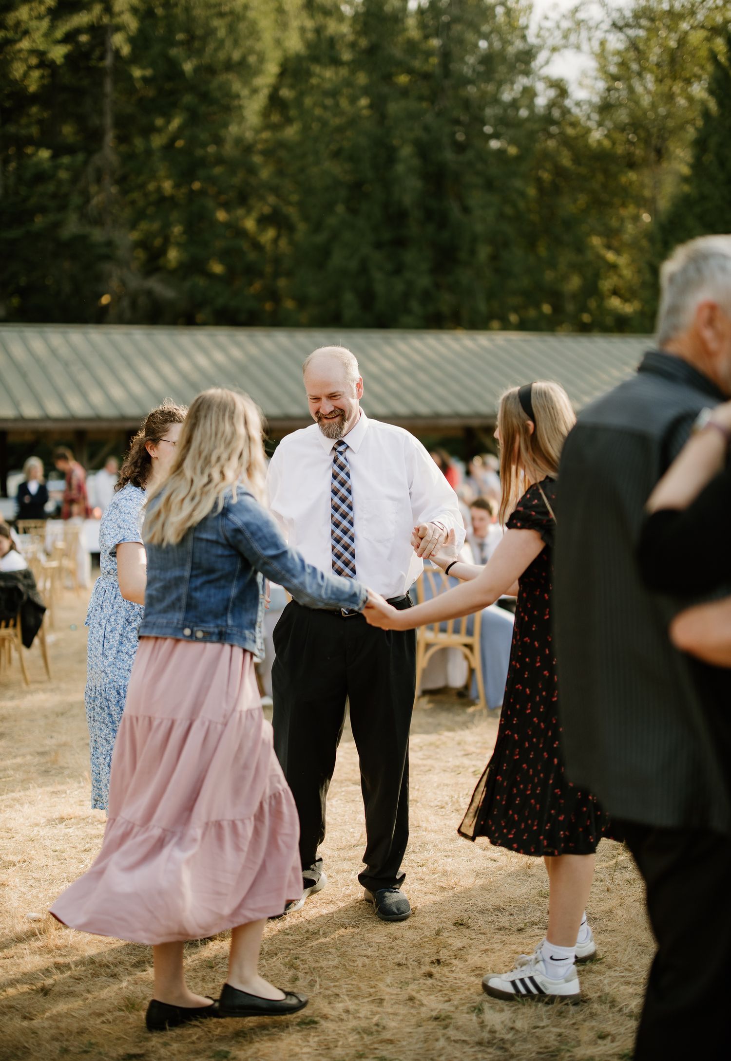 Smiling man in white shirt dances with two women in pink and black dresses at outdoor reception.