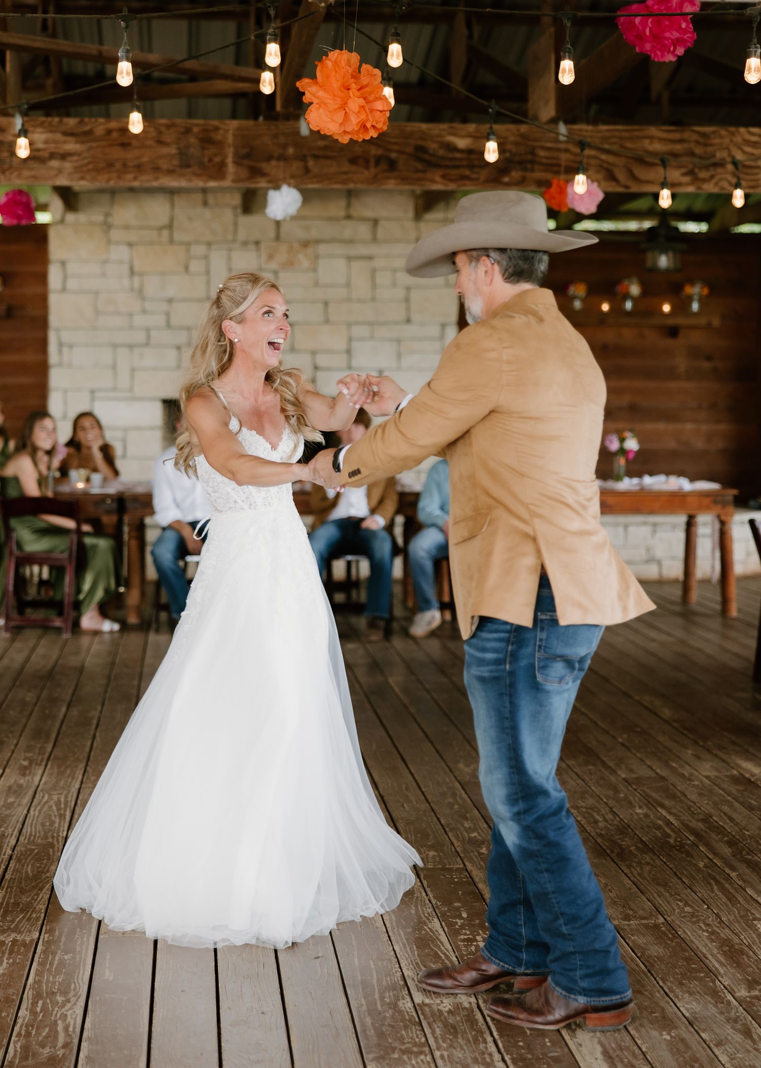 A bride in a white gown dances with her groom in a cowboy hat and tan blazer during their first dance.